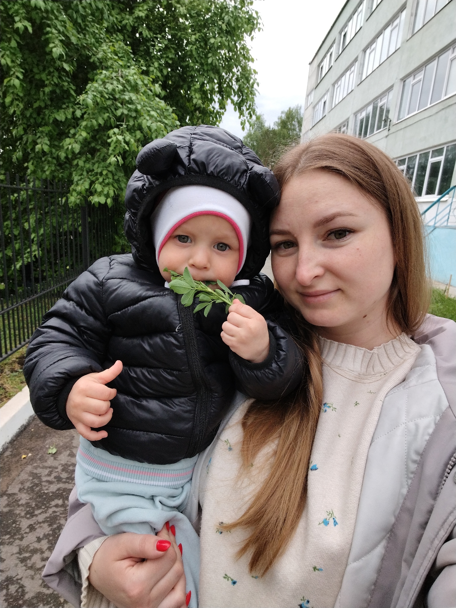 A photograph depicts a woman holding a young child outdoors. The woman is fair-skinned with light brown, shoulder-length hair, wearing a light-colored sweater with small floral patterns, and a grey quilted jacket. She looks towards the camera with a slight smile. The child, held in the woman's arms, is fair-skinned with light eyes, wearing a white hat with a red trim under a black puffer jacket with a hood. The child is holding a sprig of green leaves to their mouth.
The background includes greenery, a black metal fence, and a building with multiple windows. The location is identified as Chaikovsky, Russia.