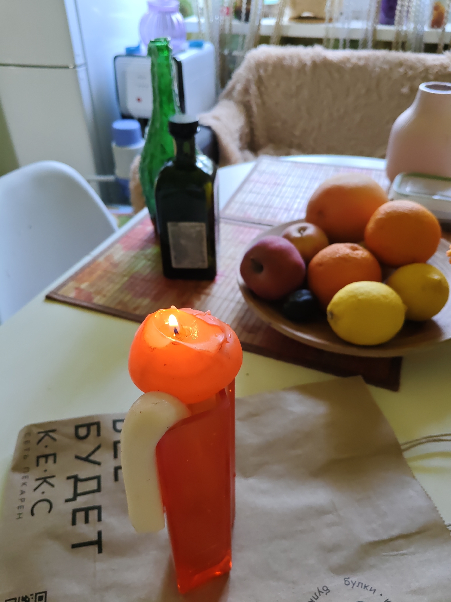 The image shows a table setting with various objects. In the foreground, a lit orange candle is placed in front of a red translucent rectangular object. To the left of the candle, there is a piece of white material. Below the candle and to the left, a piece of paper with Russian text is visible. The text includes "КЕ-КС" and "БУДЕТ", which translates to "will be". Further back on the table, a wooden plate contains several pieces of fruit, including oranges, lemons, and an apple. Behind the fruit plate, there is a dark green bottle with a black cap, and a taller, slender green glass bottle. In the background, a beige fuzzy blanket is draped over a piece of furniture, and a light pink vase is visible. A white appliance, possibly a refrigerator, is partially visible on the far left. The lighting suggests an indoor setting, likely in a domestic environment. There is no indication of the location being Rostov-on-Don, Russia, beyond the Russian text on the paper.