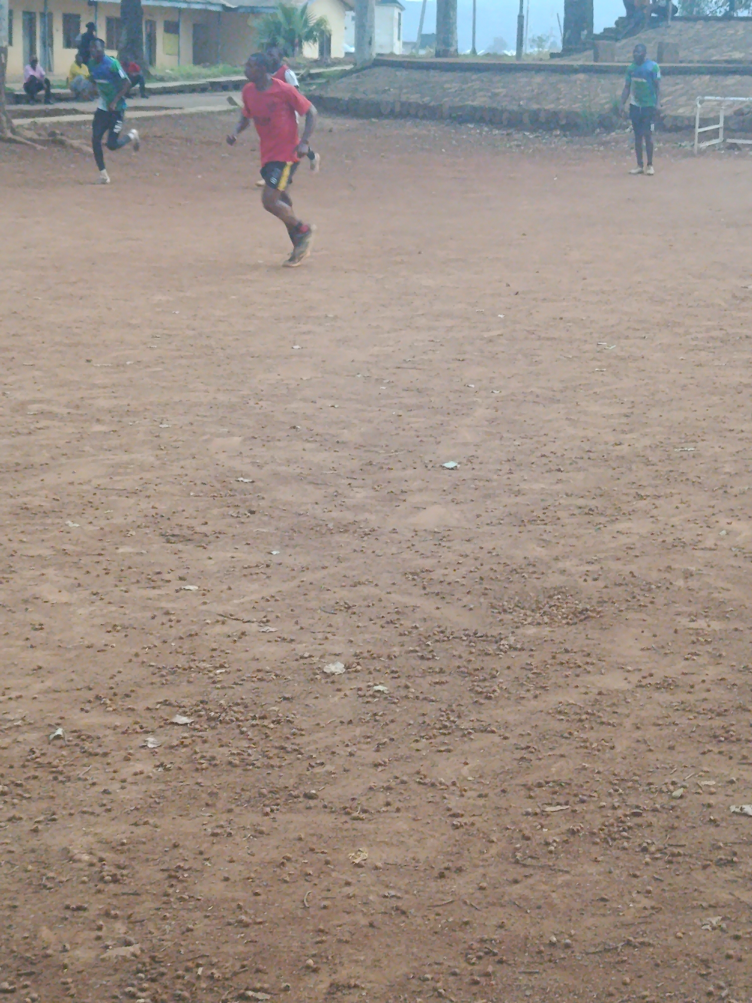 The image depicts an outdoor scene, likely a sports field, in Rafin Par, Nigeria. In the foreground, a person in a red shirt and black shorts with yellow accents is captured mid-stride, running. Behind them, another individual in a blue and green shirt and black pants is also running. Further in the background, several other people are visible, some sitting and some standing, near a low building. The ground is covered in dirt and small pebbles, and there is a white structure, possibly a goalpost, visible on the right side of the frame. The overall impression is of a casual sporting activity or training session taking place in a somewhat undeveloped outdoor area.
