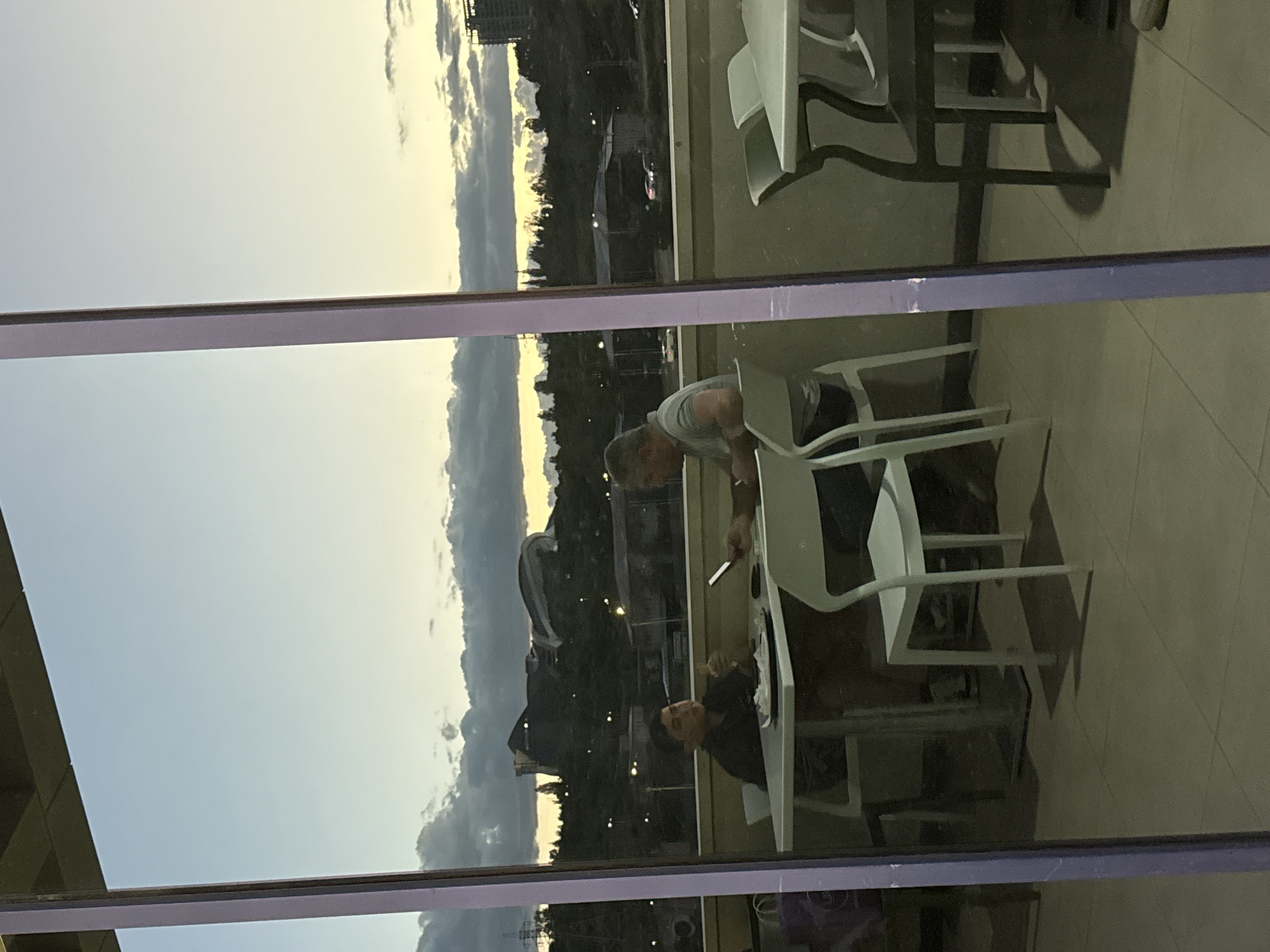 This image, set in Yerevan, Armenia, is a window reflection capturing both an indoor scene and an outdoor twilight landscape. Two men are visible in the reflection, seated at white tables indoors. The man on the left wears a dark shirt and appears to be eating or holding food, looking towards the right. The man on the right wears a light-colored shirt, looking towards the left, and holds a small, rectangular object, possibly a phone or a knife. White chairs and the tiled floor of the indoor space are also reflected.
Outside the window, a dark, cloudy sky dominates, with a warm, yellowish glow visible on the horizon, indicative of dusk or twilight. A distant cityscape with buildings and trees is silhouetted against the sky. A prominent purple-hued structural beam or window frame runs horizontally across the middle of the image. No visible text is present. The overall scene conveys a relaxed, early evening atmosphere in an urban setting.