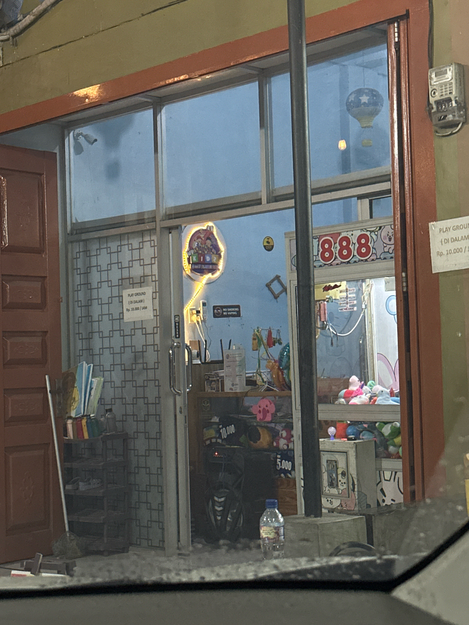 This image depicts the exterior of a building in the city of Binjai, Indonesia, likely a small entertainment venue or game center. The facade features a large glass storefront with a reddish-brown frame. A brown wooden door is visible on the left side.
Inside the building, through the glass, several elements are discernible. A sign in Indonesian reading "PLAY GROUND (DI DALAM) Rp. 10.000/JAM" indicates a playground area with an hourly fee. A neon sign with cartoon characters and the text "PLAY KIDZ" is illuminated. In the center, a claw machine filled with plush toys is visible, topped with a sign displaying "888".
Further inside, shelves are stocked with various items, including what appear to be books or magazines near the entrance and a collection of plush toys within the claw machine and on display. A motorcycle is parked to the right of the claw machine, and a plastic water bottle rests on a concrete block in the foreground.
The environment suggests an indoor play area or arcade, possibly at dusk or evening, given the lighting. A detached electrical meter is mounted on the exterior wall to the right of the storefront.