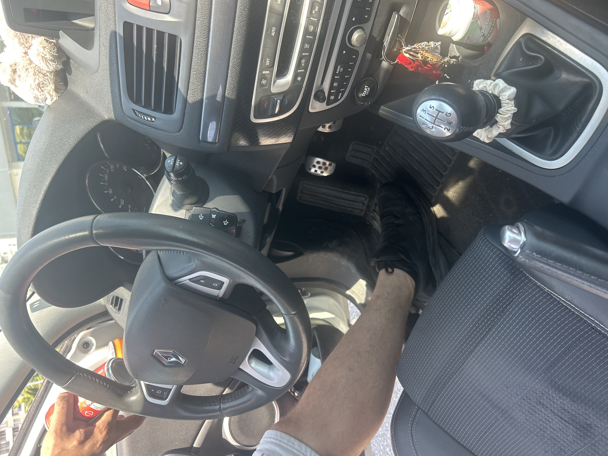 In Plovdiv, Bulgaria, inside a car, a person's right leg and arm are visible. The person's leg is extended and their foot, clad in a dark sneaker, is pressing down on the clutch pedal. The arm, holding a red-and-white can, is positioned near the steering wheel, suggesting the person might be either driving or preparing to do so. The car's interior, dominated by black and gray tones, features a steering wheel with visible controls, a dashboard with a speedometer displaying 150 km/h, a manual gear stick with a white scrunchie around its base, and a center console holding keys and a packet of chewing gum. The overall scene suggests a moment of pause within the vehicle, possibly before or after a journey.