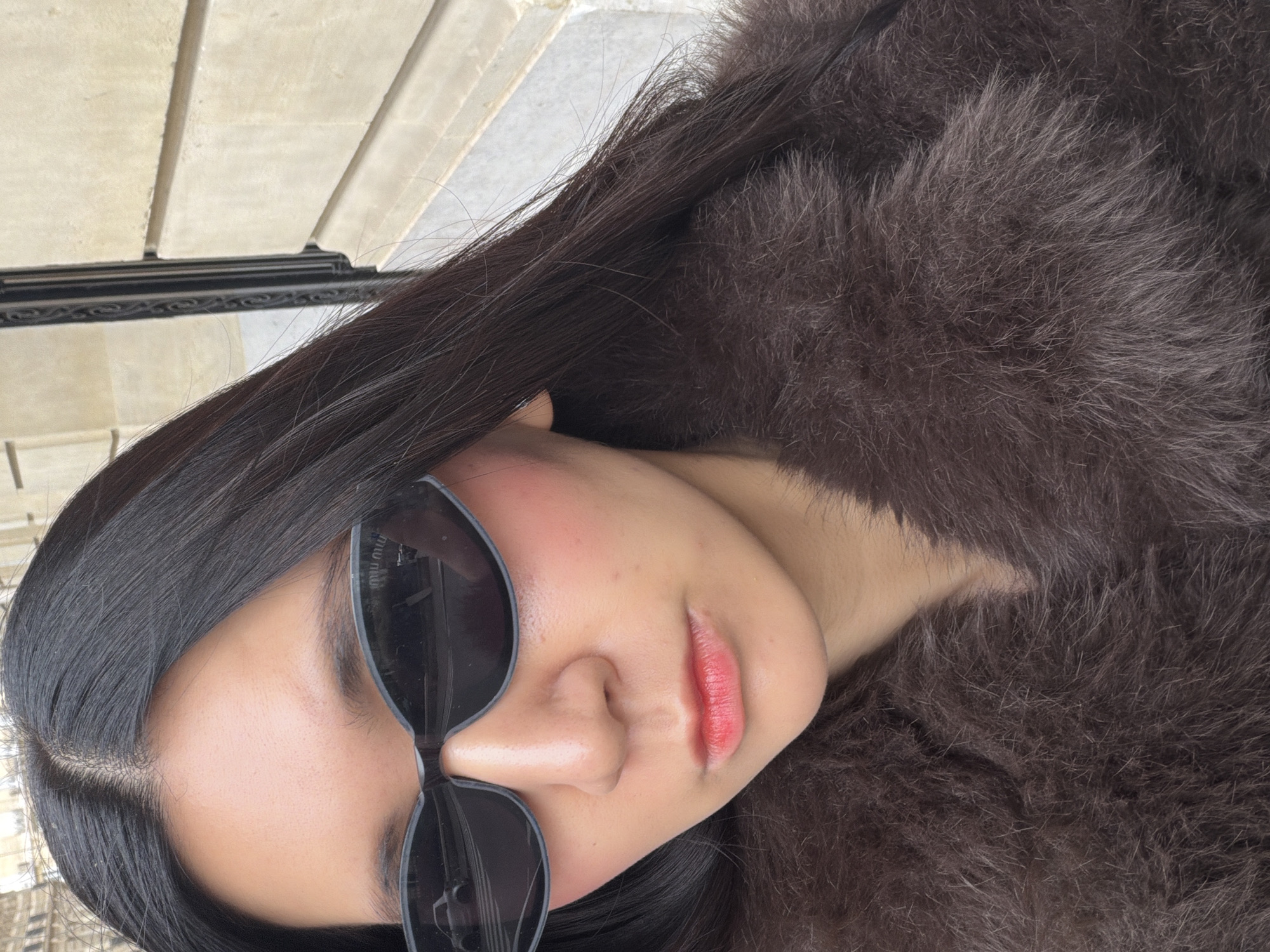 A person wearing sunglasses and a fur coat poses in Paris, France. The person has dark hair and is wearing red lipstick. The background includes architectural details consistent with Parisian buildings.