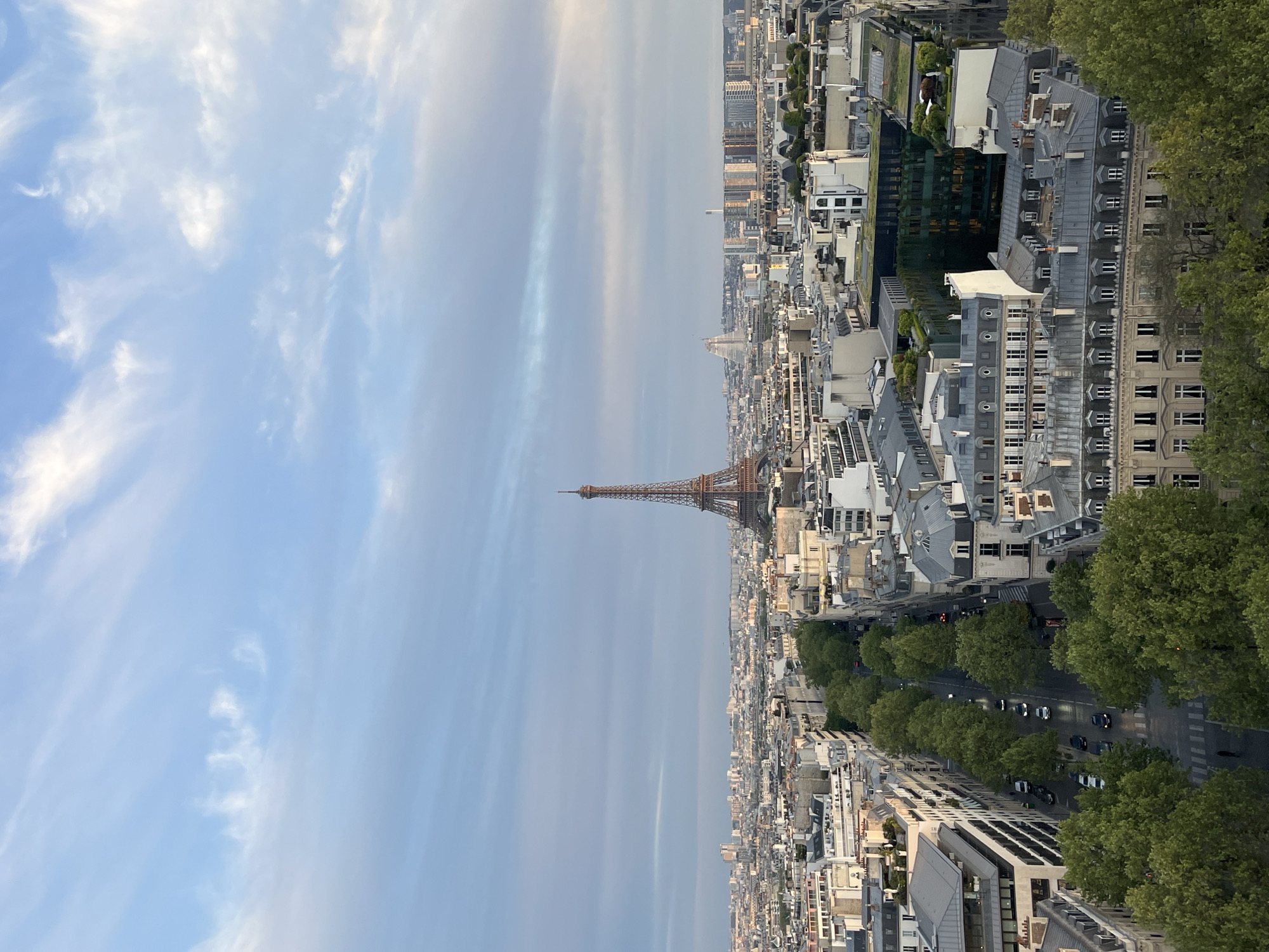 The image displays a high-vantage point, panoramic view of the Parisian cityscape. The iconic Eiffel Tower stands prominently on the right side of the frame, reaching towards the sky. The sky, occupying the upper portion of the image, is a light blue with scattered, wispy white clouds.
Below the sky, the city stretches across the entire frame, showcasing dense urban development. Numerous buildings with classic Parisian architecture, characterized by their grey, mansard roofs and ornate facades, fill the midground and foreground. A wide, tree-lined avenue, adorned with lush green trees, winds through the city in the lower-middle section of the image. Several cars are visible on this street, indicating ongoing traffic.
The scene captures a typical daytime setting, likely during morning or late afternoon, given the soft, diffused natural light and the absence of harsh shadows. The weather appears fair with light cloud cover. The overall impression is one of an expansive, historic, and beautiful metropolis. There is no visible text in the image.