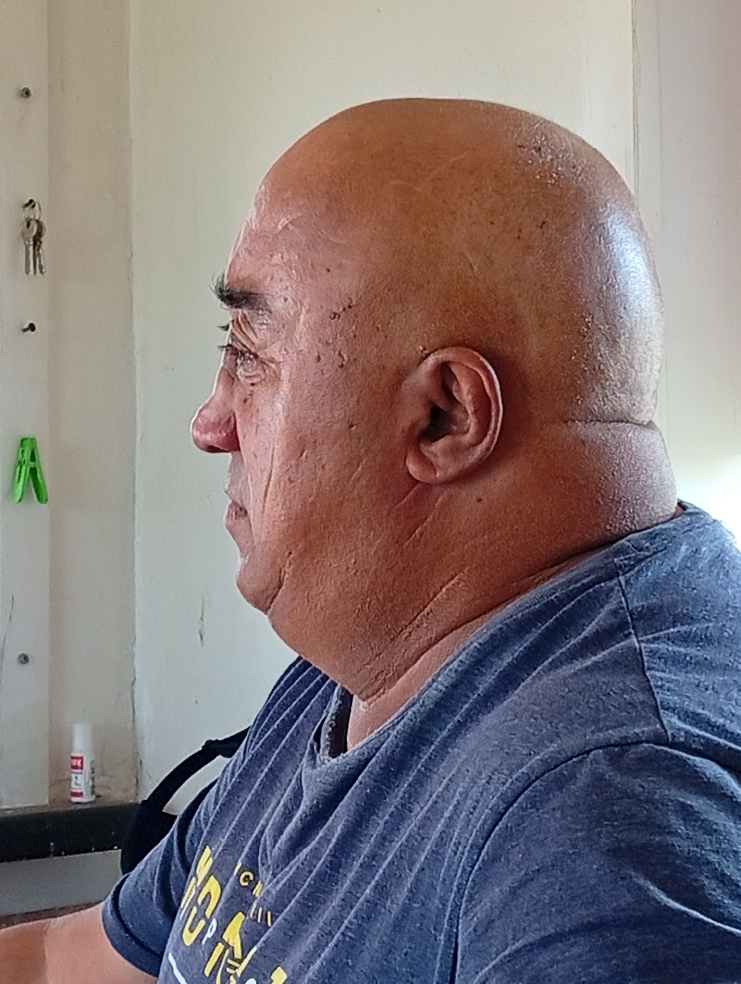 A medium close-up profile shot shows a bald, middle-aged man with tanned skin and wrinkles. He is wearing a dark blue t-shirt. The man is positioned on the right side of the frame, with his head and shoulders filling much of the space. His face is turned to the left, and his expression is neutral. The background is a plain, light-colored wall, with a few objects visible. To the left of the man, on the wall, are two keys hanging on a hook, and a green clothespin. Further down, on a shelf, there is a small white bottle with red writing. The scene appears to be indoors, possibly in a home or office setting in Қызылжар ауылдық округі, Kazakhstan.