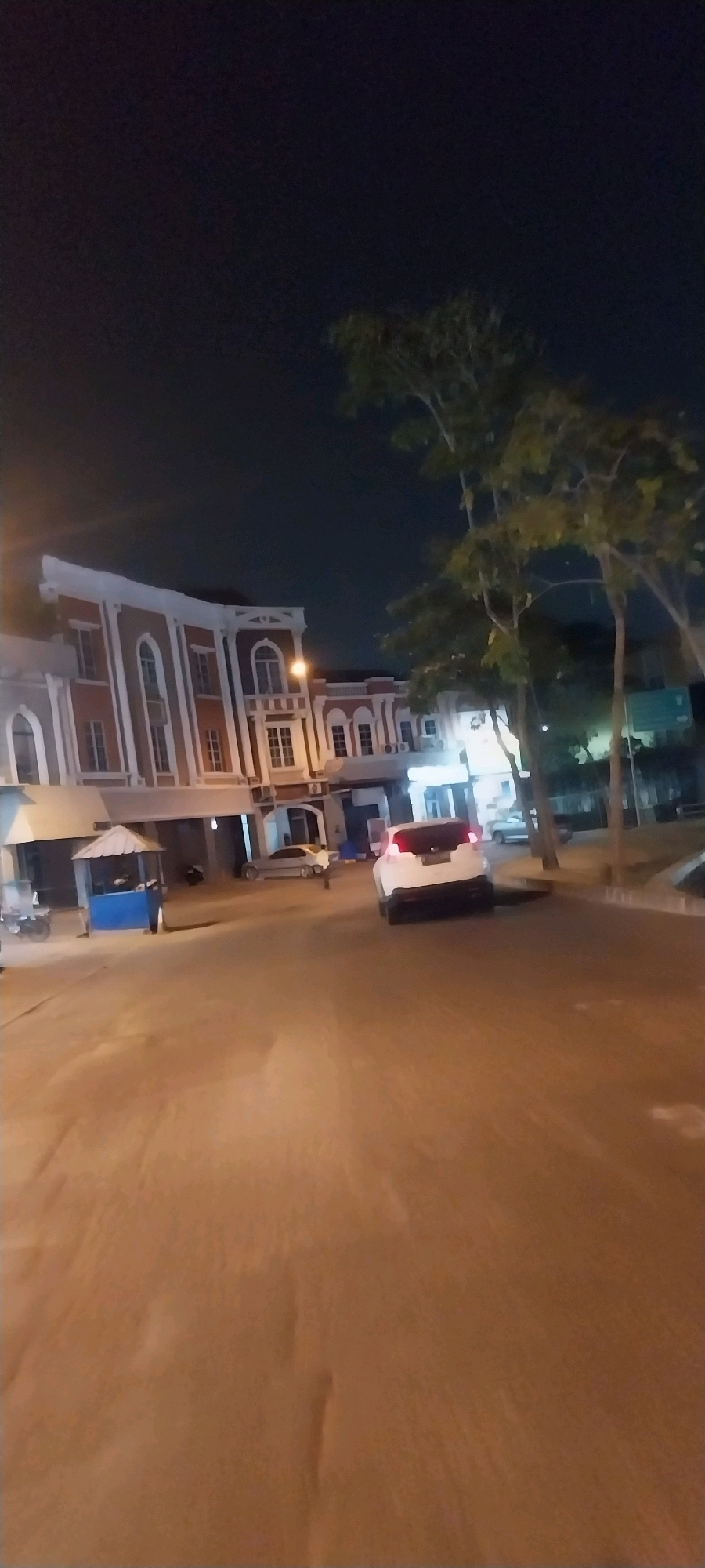This is a low-light, slightly blurry, vertical-orientation photo of a street scene at night in Kelapa Dua, Indonesia. A two-story building with a reddish-brown facade and white trim, featuring arched windows, is visible on the left. In front of the building, there is a small blue kiosk or shelter with a white roof. A white car is parked on the right side of the street, facing away from the camera. Trees with sparse foliage are present on the right side of the image, with some lights from other buildings visible in the background. The street surface appears to be paved and reflects some of the ambient light. The sky is dark and clear.