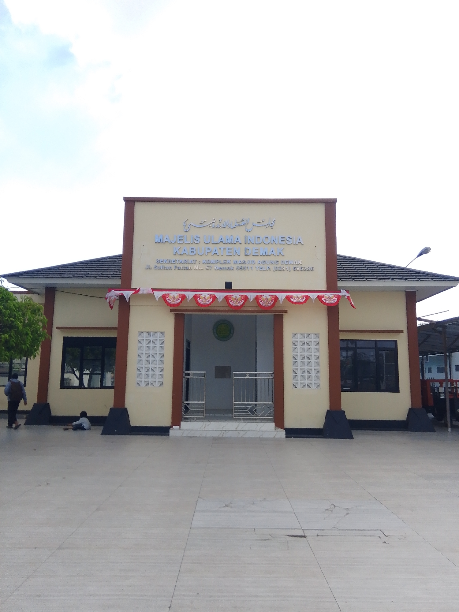 The image shows the front of a building with signage that reads "MAJELIS ULAMA INDONESIA KABUPATEN DEMAK". Below this, it states "SEKRETARIAT: KOMPLEK MASJID AGUNG DEMAK" and an address "JL. SULTAN PANGLIMA NO. 57 DEMAK 59511 TELP (0291) 666396". The building is cream-colored with brown trim and has a dark-tiled roof. In front of the building, there is a paved area with decorative flags strung across the entrance. Two individuals are visible: one person walking away from the camera on the left side of the frame, and another person sitting on the ground in front of the building. The environment appears to be an outdoor setting with a tree on the left and a street light on the right. The sky is overcast with visible clouds.