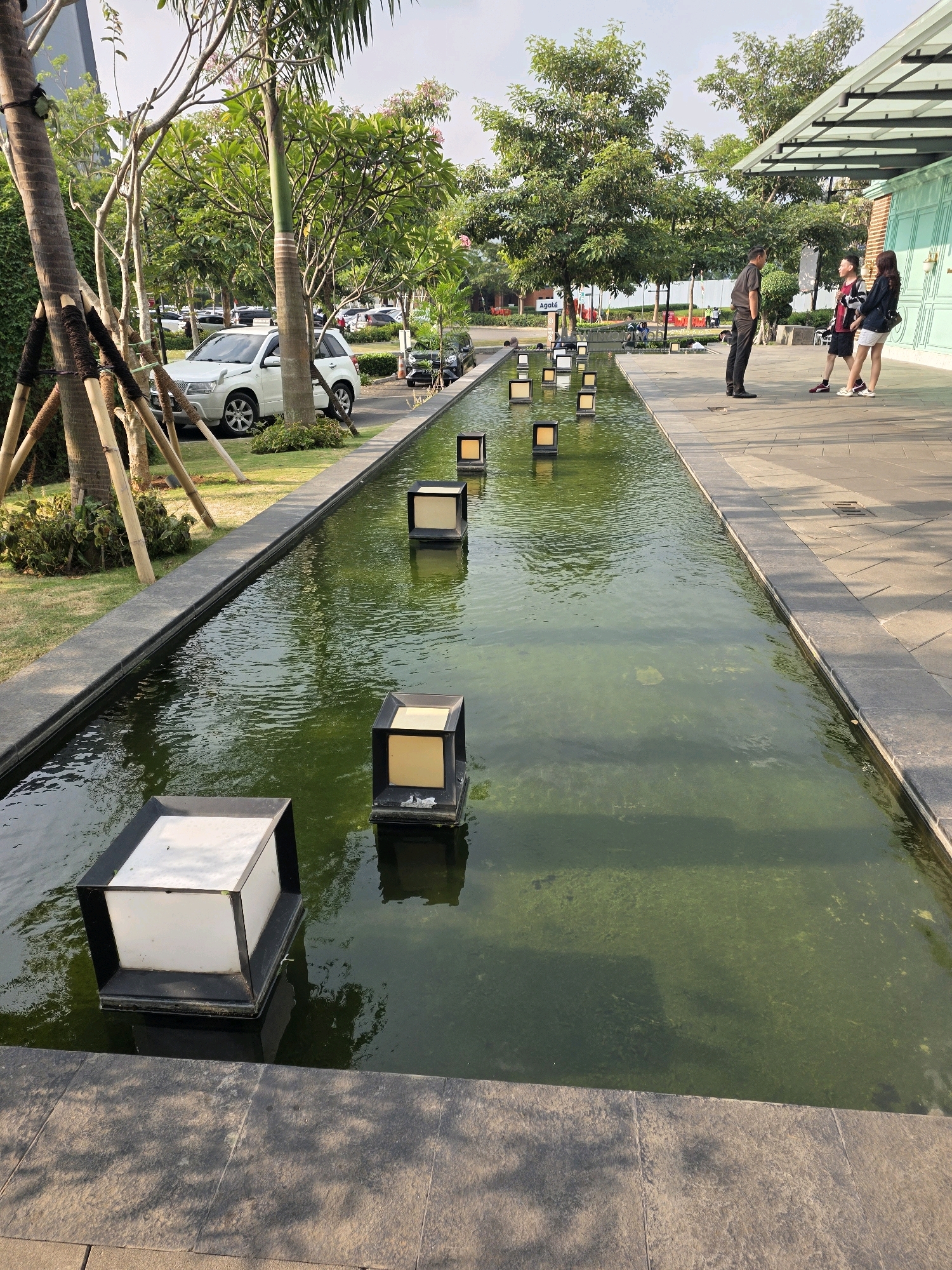 The image shows a long, narrow outdoor fountain with several submerged cube-shaped lights. The water in the fountain is a murky green color, with algae visible on the bottom in some areas. The fountain is surrounded by a paved walkway and a grassy area with trees. In the background, there are several cars parked and a building with large windows.
Three people are visible on the walkway to the right of the fountain. One person, a male wearing a dark shirt and pants, is facing towards two other individuals. The two other individuals are a male and a female, both dressed casually. They appear to be engaged in conversation.
The overall environment suggests a public space, possibly a park, a plaza, or an area adjacent to a commercial establishment. The presence of cars indicates it is likely in an urban or semi-urban setting.
The provided crops highlight specific elements:
- The first crop focuses on the vegetation and a white SUV parked near the fountain.
- The second crop shows the three individuals on the walkway and the building's exterior.
- The third and fifth crops provide a closer view of the fountain lights and the water.
- The fourth and sixth crops show the paved walkway and the green water of the fountain.
Based on the architectural style and the general ambiance, this scene could potentially be located in a modern commercial or recreational area within Bandung City, Indonesia. However, without explicit signage or more distinctive landmarks, pinpointing the exact location within Bandung is not possible from the image alone.