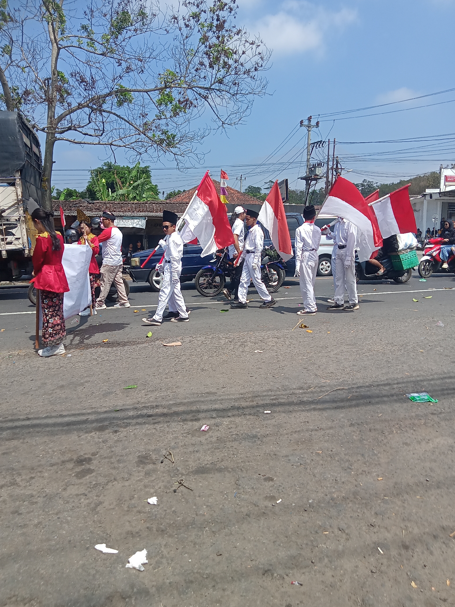 In Urutsewu, Indonesia, a parade is underway on a paved road under a clear blue sky. The parade features several individuals, some of whom are wearing white uniforms and hats, marching and holding Indonesian flags. One individual in the foreground on the left is wearing a traditional red and gold outfit with a patterned skirt. Another person in the center is wearing a red and white shirt and light-colored pants. In the background, there are houses with tiled roofs, trees, utility poles with electrical wires, and parked vehicles, including a truck on the left and motorcycles. The road appears to be somewhat worn and has scattered debris, including paper and plastic. Some of the parade participants are walking next to or behind motorcycles.