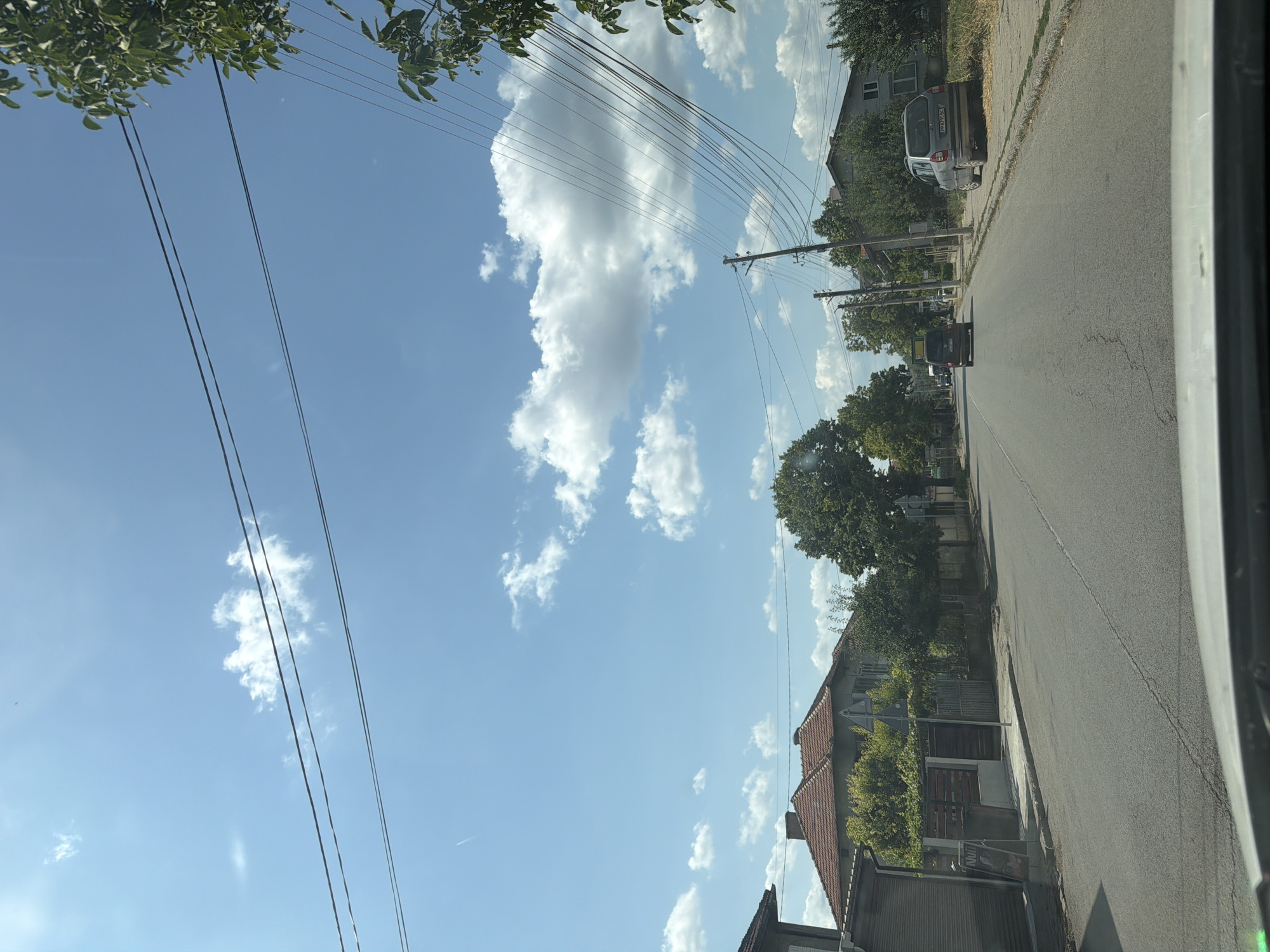 The image captures a street scene in Tsar Kaloyan, Bulgaria, under a bright blue sky with scattered white clouds. The perspective appears to be from inside a vehicle, looking forward down a paved road. On the left side of the road, there are houses with red tiled roofs and some trees. A building with a gray roller door is visible closer to the foreground. A street sign is also present on the left.
The road itself is asphalt with visible cracks. Two cars are on the road, one ahead of the other, both moving in the same direction. The first car, a dark minivan, is further down the road. The second car, a silver hatchback, is parked on the right side of the road, slightly ahead of the viewpoint.
On the right side of the road, there is a sidewalk and more trees. Utility poles with numerous wires are prominently visible, stretching across the frame and contributing to the overhead visual clutter. Some of the wires are draped low over the road. The overall impression is a typical residential street in a Bulgarian town on a sunny day.