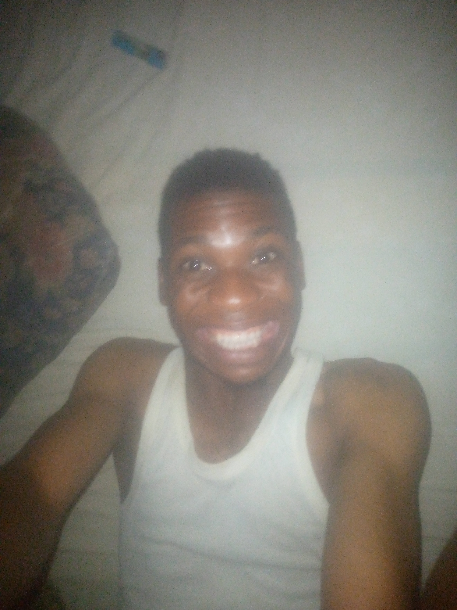 A person is lying in bed. The person is wearing a white tank top and is smiling. The background is blurry.