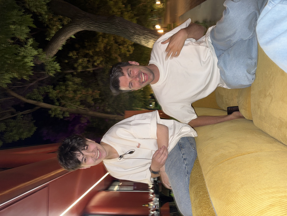 Two people sit on a yellow corduroy sofa. The person on the left wears a light-colored t-shirt and jeans, smiling. The person on the right is sitting further back, wearing a white t-shirt and jeans, grinning widely. A phone is visible between them on the sofa. A reddish-brown structure is partly visible on the left, with dark foliage and lights overhead.