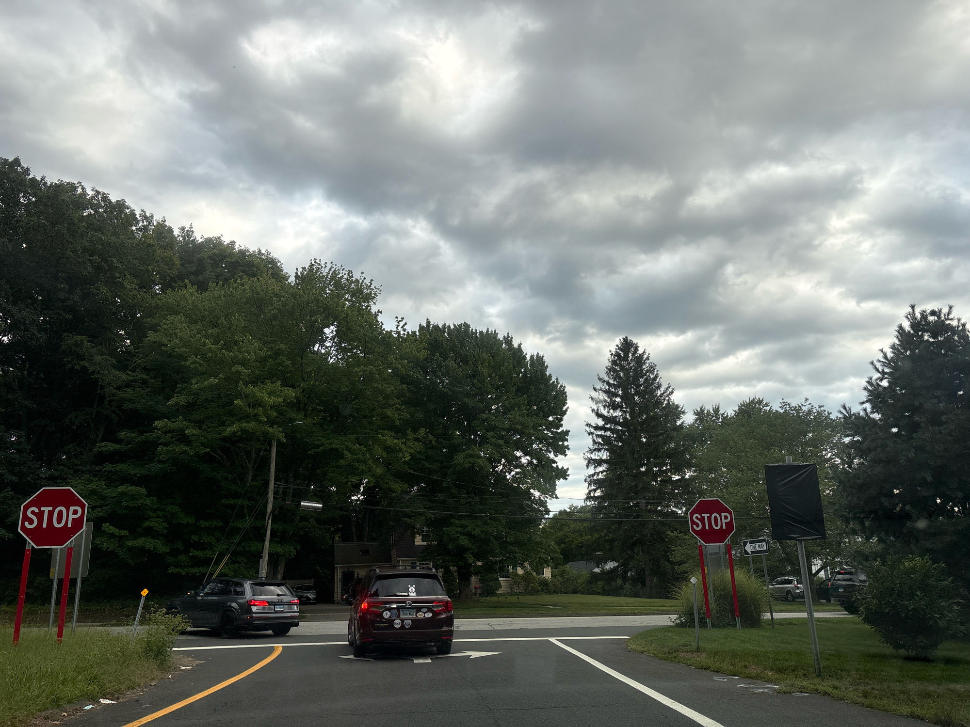The image shows a street scene in Glastonbury, United States of America. In the foreground, two cars are visible on a paved road with yellow lane markings. The car on the left is a dark-colored SUV, and the car on the right is a maroon minivan with several stickers on its rear. Both cars are stopped at an intersection.
To the left of the intersection, there is a red stop sign and a sign indicating "ONE WAY." Tall trees with green foliage line the sides of the road. The sky is overcast with gray clouds. In the background, houses and more trees are visible. There are no people clearly visible in the image. The scene appears to be a typical suburban intersection.