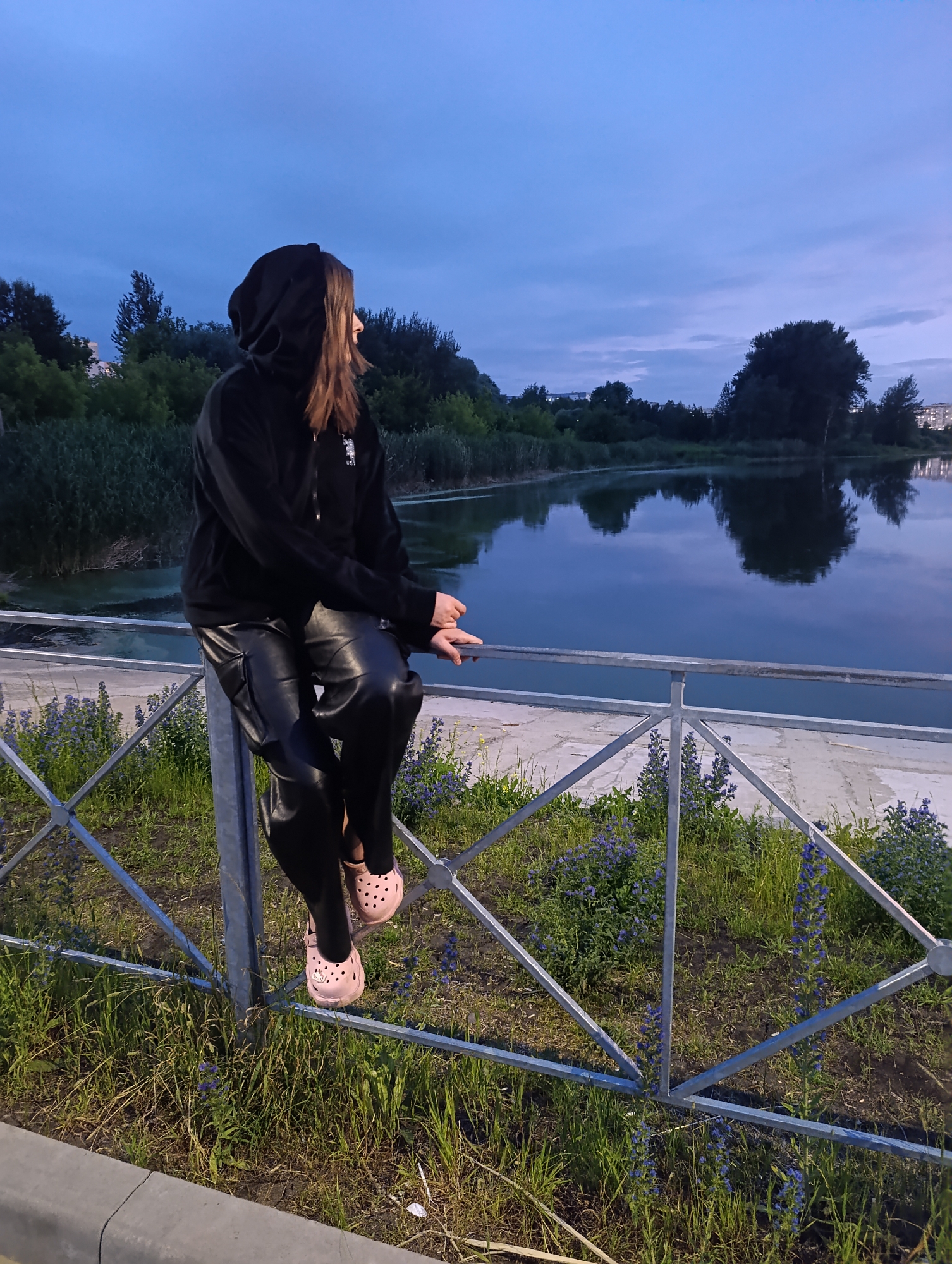 The image shows a person sitting on a metal fence with a body of water behind them. The person is wearing a black hooded sweatshirt and black pants, and pink clogs. The water is calm and reflects the trees and sky. The sky is a deep blue with some clouds. There are trees and bushes on the far side of the water, and some buildings in the distance. The scene appears to be a park or a waterfront area. Given the provided metadata, this image was taken in Penza, Russia.