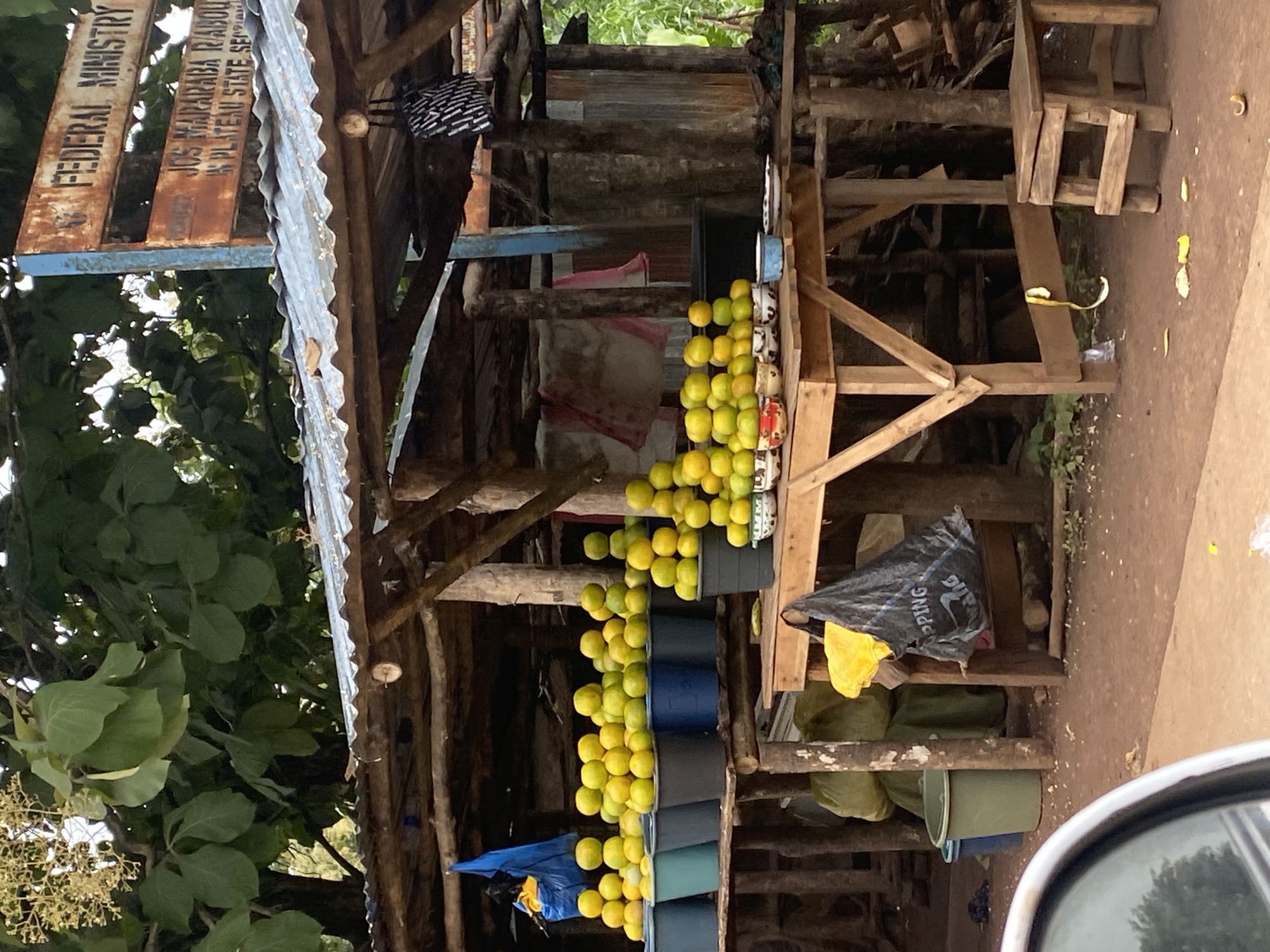 The image shows an outdoor fruit stall with a variety of oranges displayed in buckets and bowls. The stall has a corrugated metal roof supported by wooden beams. A signpost with "FEDERAL MINISTRY" and other text is visible in the upper right corner. In the background, trees and foliage are present. The ground is dirt, with some scattered debris. Several plastic buckets and bags are also visible around the stall. The scene appears to be in a rural or semi-urban setting.