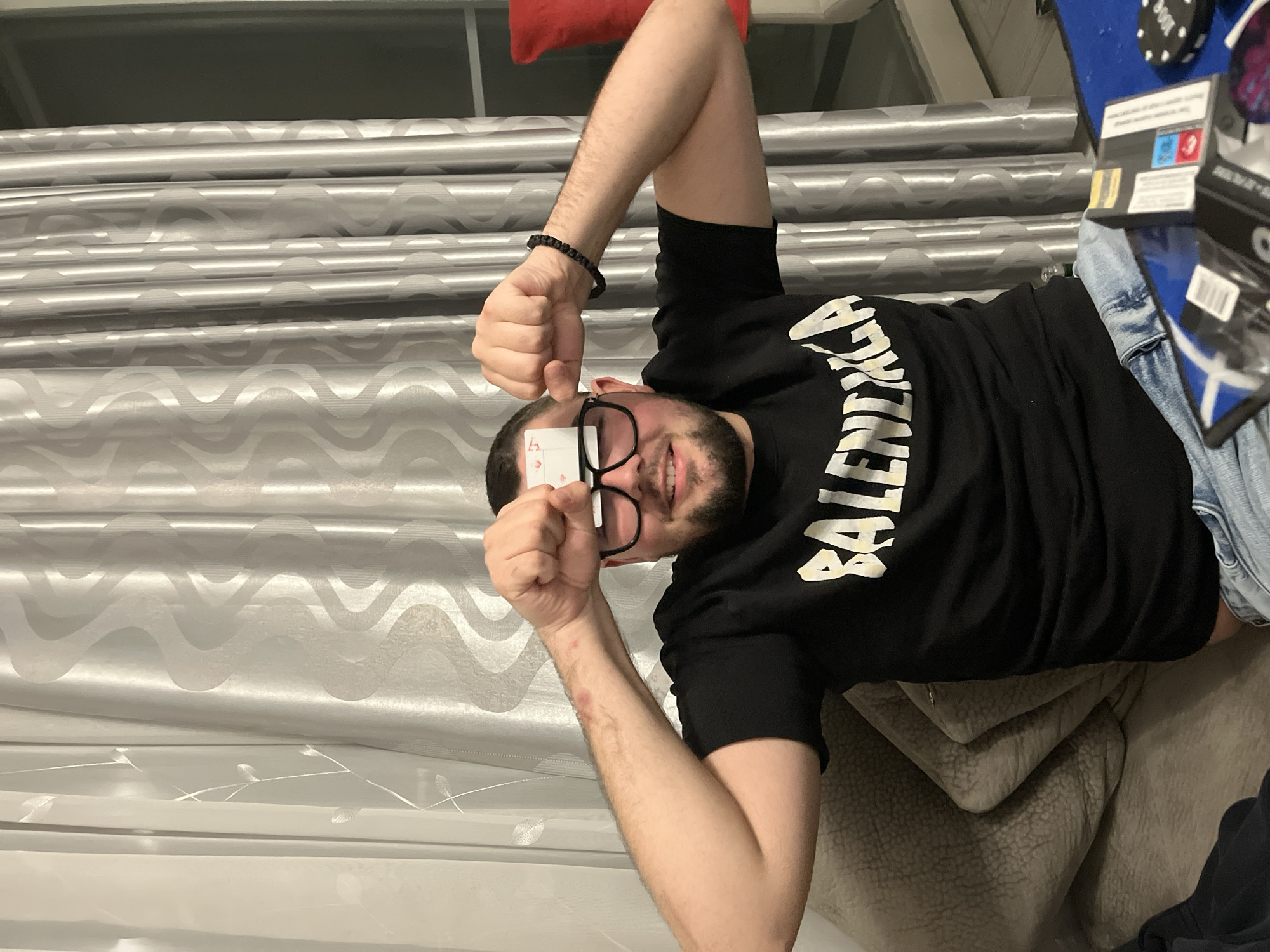A man with a beard and glasses is holding a playing card to his forehead. He is wearing a black Balenciaga T-shirt and a black bracelet. His arms are raised and bent at the elbows, and his fists are clenched. He is reclining on a light brown sofa in front of a window with silvery-white curtains. A red pillow is visible above his head. Other items, including what looks like a game box, are visible on the sofa next to him. The specific location in the city of Burgas, Bulgaria is not discernible from the provided visual information.