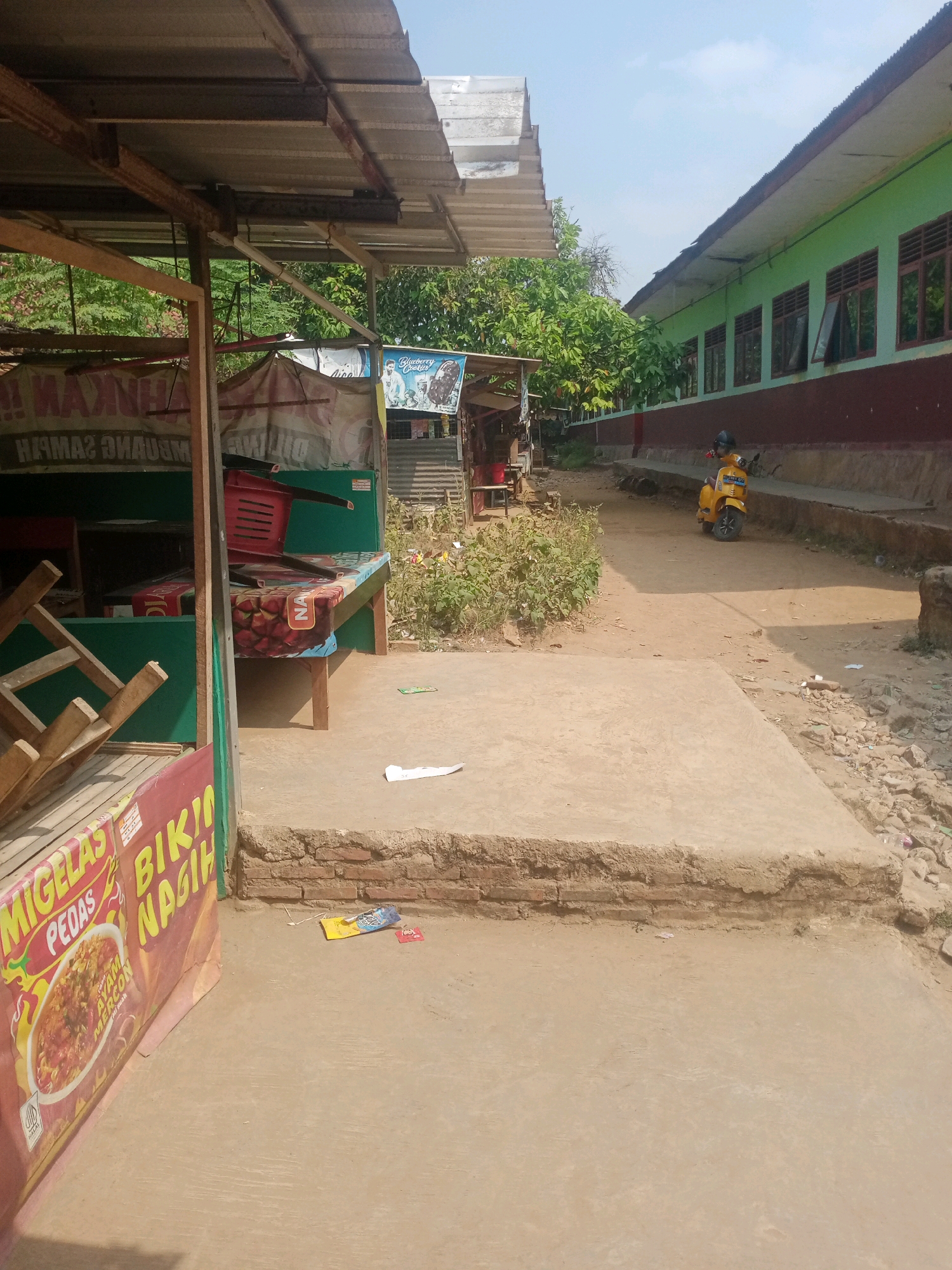 The image shows an outdoor scene in what appears to be a rural or semi-urban area. In the foreground, a food stall is partially visible on the left, with a sign advertising "MIGELAS PEDAS BIKIN NAGIH." The stall has a tin roof supported by metal beams and wooden poles. Inside the stall, a red chair is overturned on a table. Adjacent to the stall is a concrete pathway and a raised concrete platform with exposed brickwork at the edge.
To the right of the pathway, a yellow scooter is parked. Further down the pathway, a long, light green building with a dark red base runs alongside. The building has multiple windows, some of which are open. Trees and vegetation are visible in the background. The sky is clear and blue. The ground is a mix of dirt, gravel, and concrete. There are scattered pieces of litter on the ground.