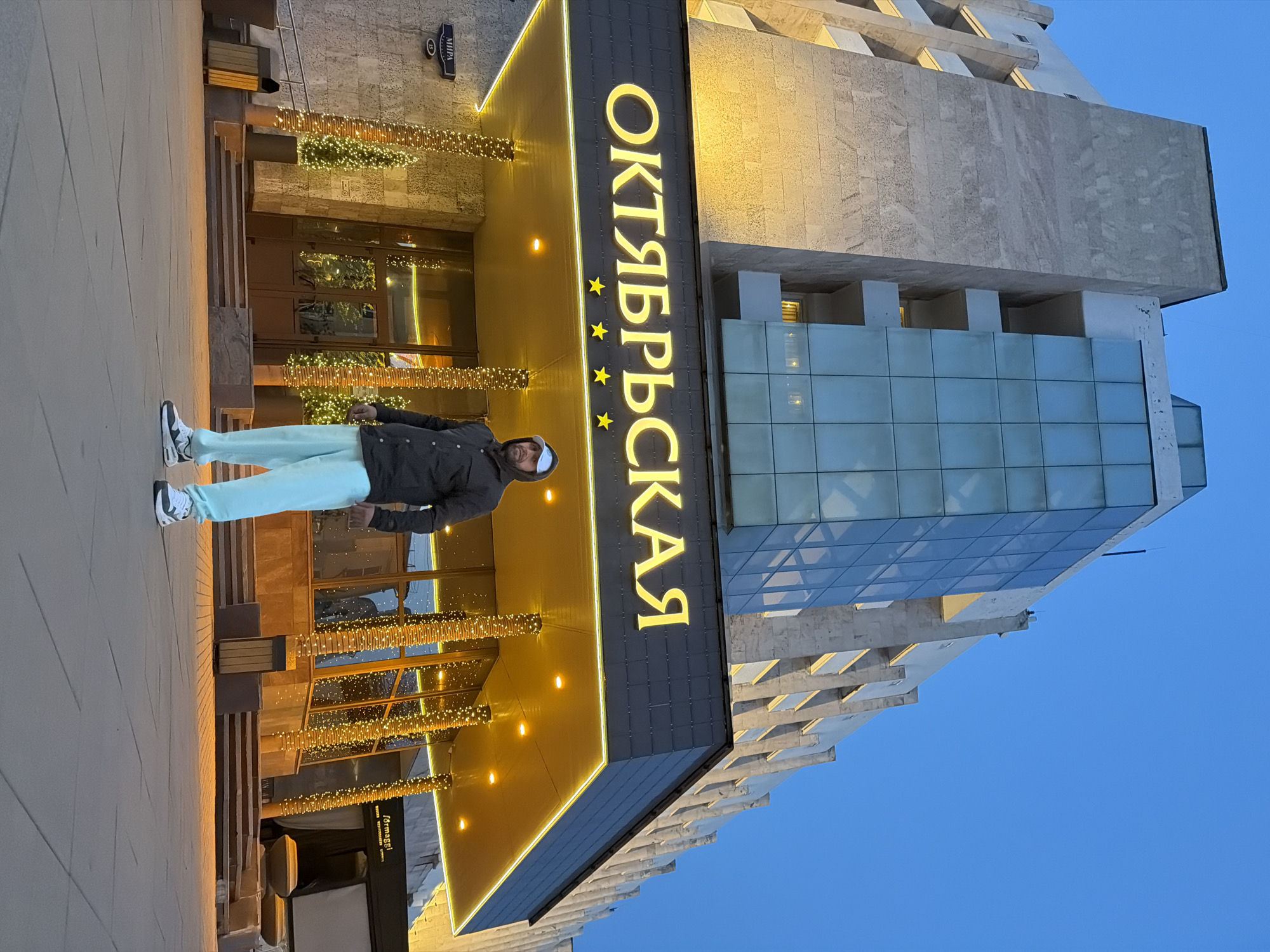 A man stands in front of the Hotel Oktyabrskaya in Krasnoyarsk, Russia. The hotel's name, "ОКТЯБРЬСКАЯ," is displayed vertically in gold letters. Decorative lights adorn the entrance. The building has a combination of stone and glass sections. The sky is blue. The man is wearing light blue pants, a dark jacket, and a white hat.