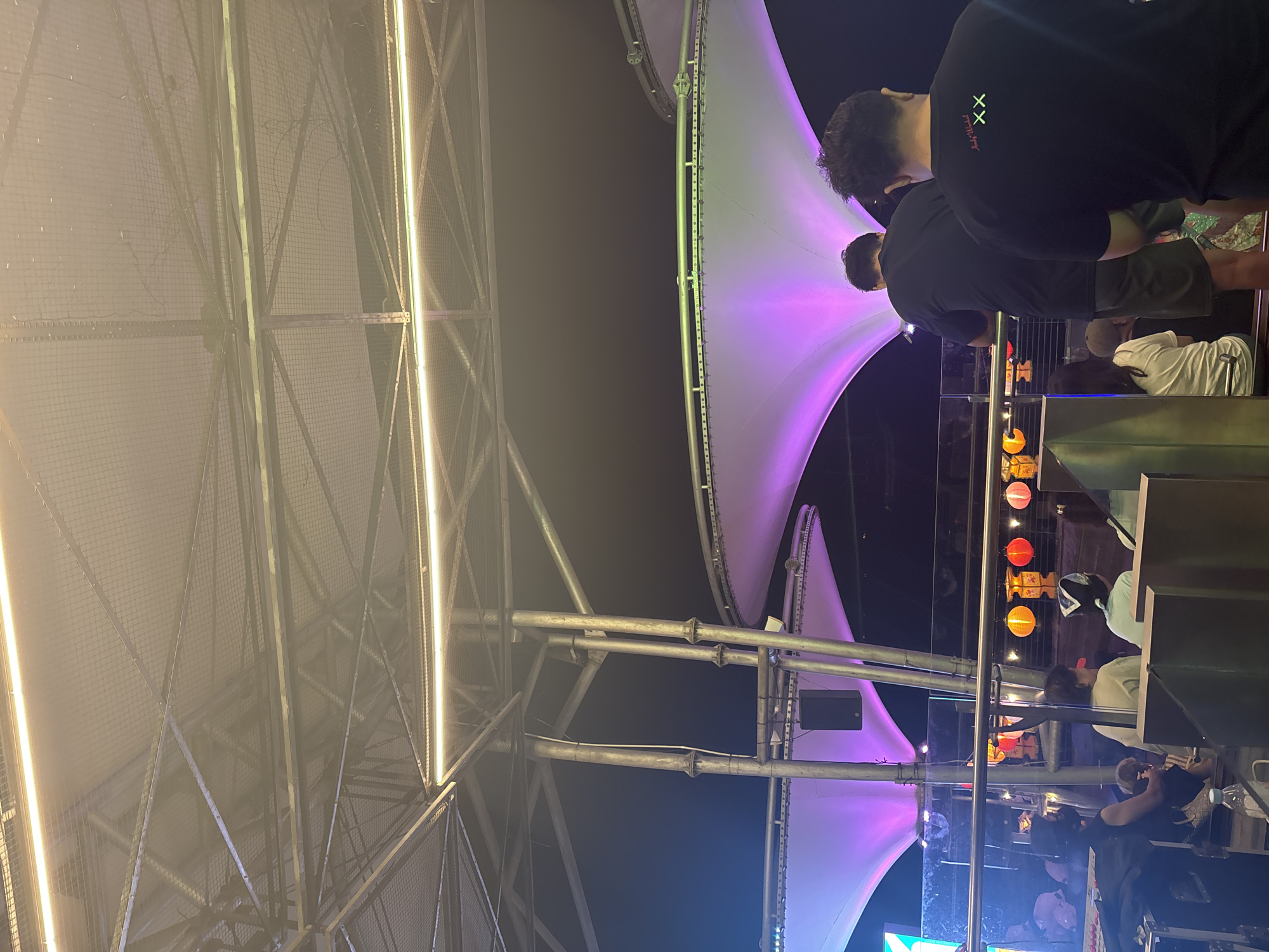 The photograph shows an outdoor scene at night. In the foreground, a metal structure with netting is partially visible, illuminated by vertical lights. The background features a large, white tent-like structure lit with purple and green lights. Two people wearing dark clothing are standing near a railing, with a third person behind them. A series of red and orange lanterns hang from the railing. Other individuals are visible behind the railing, including one wearing a white shirt and hat. The scene is located in North Jakarta, Indonesia.