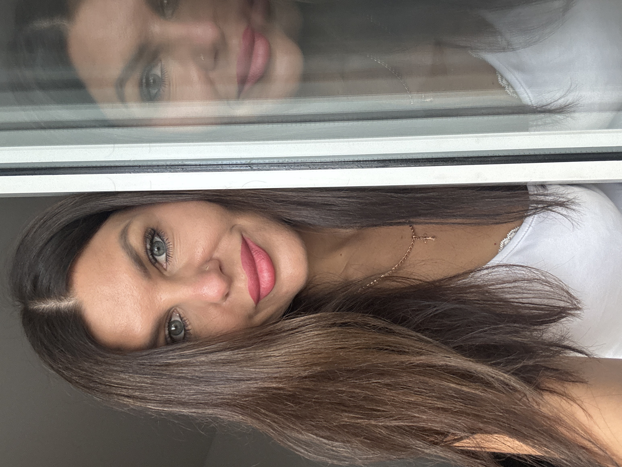 A woman with long, dark brown hair and blue-green eyes is captured in a close-up portrait, partially obscured by a window frame. She is wearing a white tank top and a delicate gold necklace with a cross pendant. Her lips are painted a vibrant pink, and she has a small nose ring. The right side of the image shows a reflection of the woman in the glass, providing a slightly distorted view of her face and upper body. The background is dark and out of focus. The scene is indoors, with the light source appearing to be from the left, casting a soft glow on her face. There are no discernible elements in the image that would indicate a specific location within the city of Chaikovsky, Russia, or any other specific urban environment.