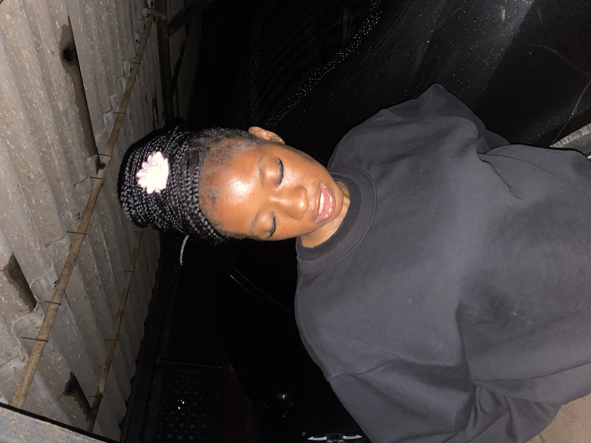 A young Black woman with her hair in braids styled in a bun adorned with a pink flower is captured from the chest up, facing downward with her eyes closed. She is wearing a black crewneck sweatshirt. Behind her, a black car is visible, its surface speckled with water droplets. The background features a corrugated metal roof structure. The scene appears to be outdoors, possibly under a shelter or carport, with a hint of what might be a tiled floor beneath. The lighting suggests it is evening or nighttime. The image was taken in Jos, Nigeria.