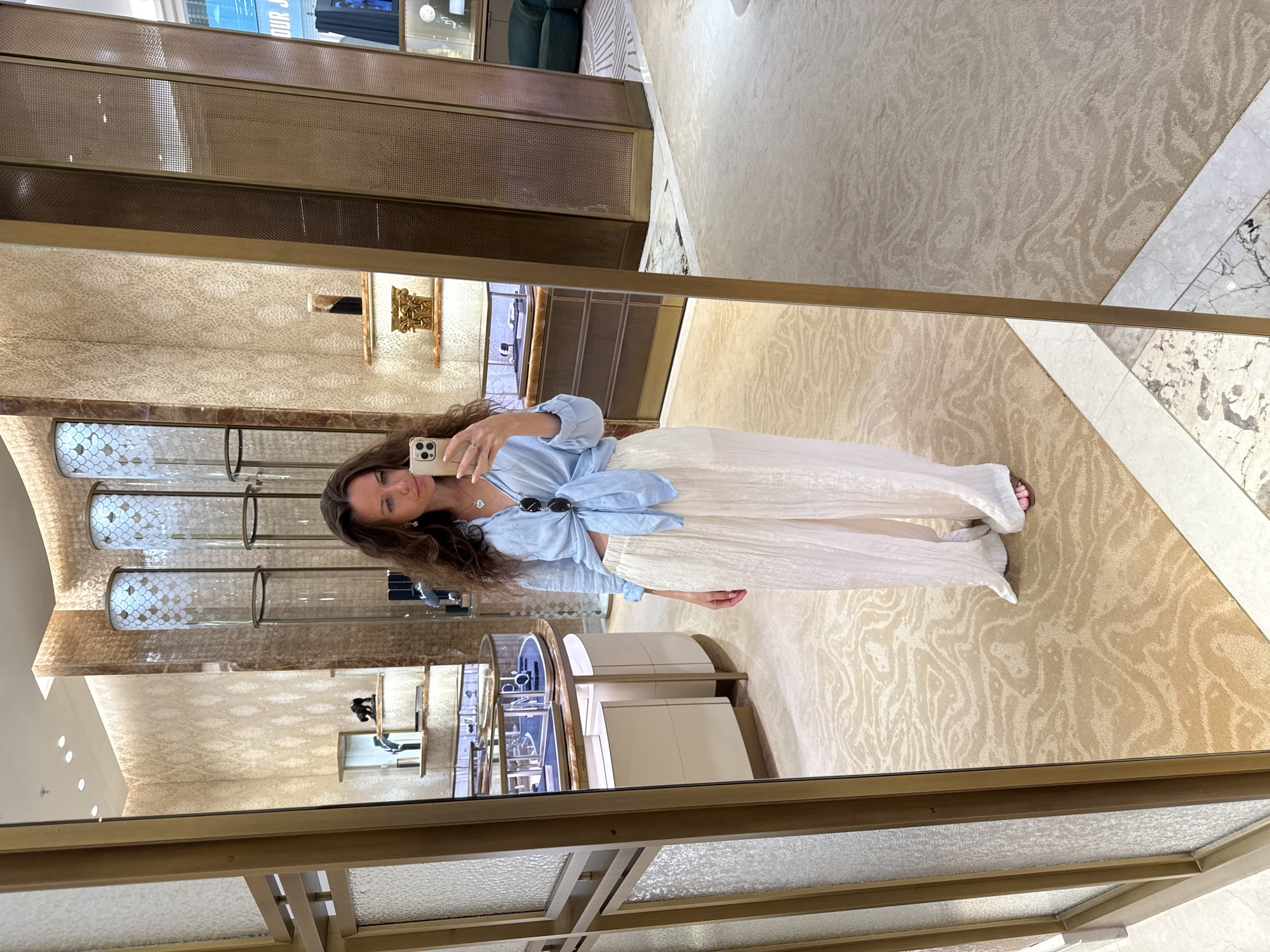 A woman is taking a mirror selfie in what appears to be a luxury boutique. She is wearing a light blue, tied-front shirt and loose, cream-colored pants. She has long, dark, wavy hair and is wearing a necklace with a blue pendant and sunglasses pushed up on her head. She is holding a smartphone in her right hand. The background shows a well-lit interior with display cases showcasing jewelry, elaborate wall textures, and textured carpeting. The overall impression is one of a high-end retail environment, possibly a jewelry store in a prominent shopping district in Dubai, United Arab Emirates, known for its luxury shopping experiences.