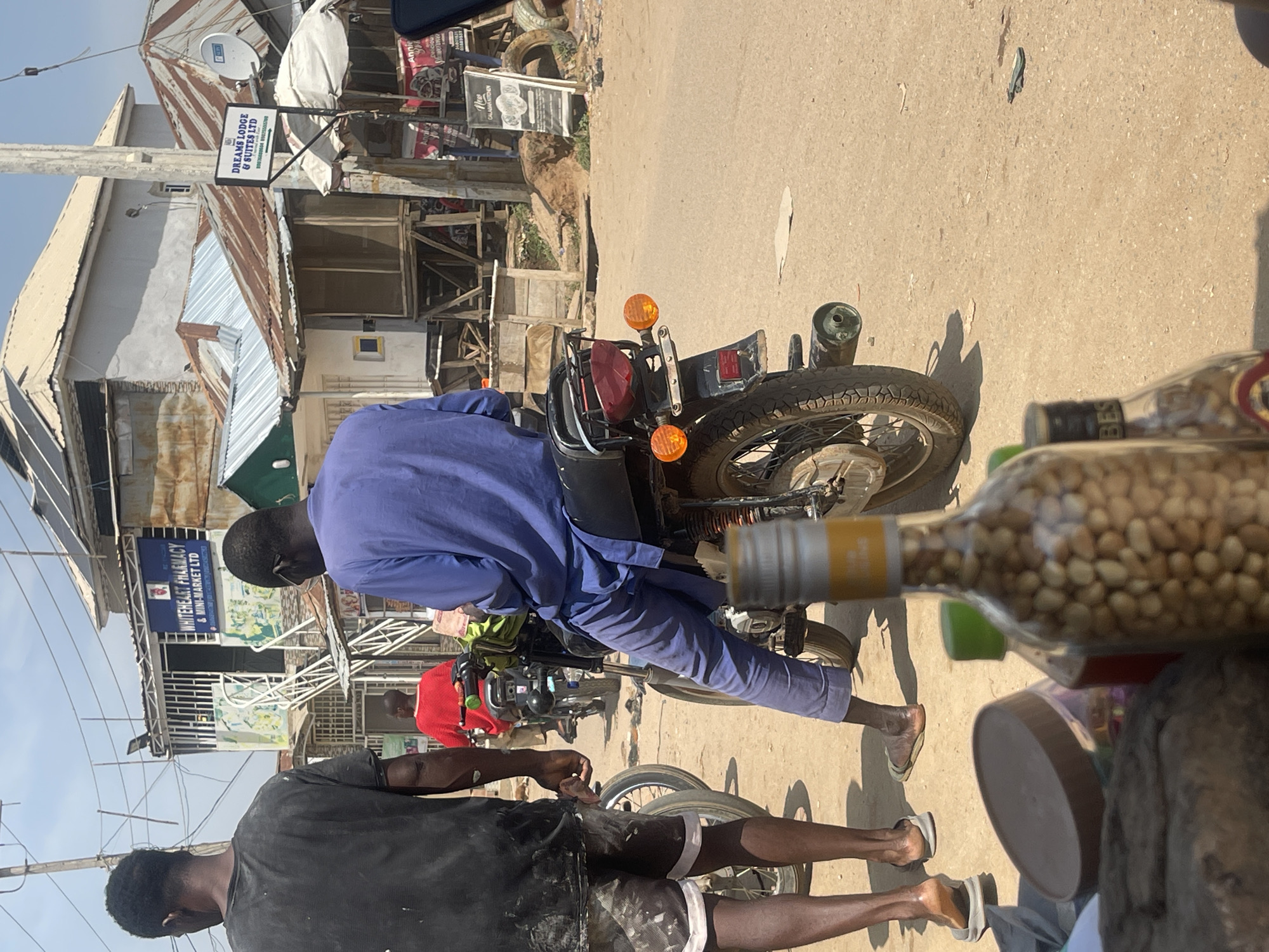 In an outdoor scene in Abuja, Nigeria, the foreground is occupied by a motorcycle, with its rear tire, exhaust pipe, and taillight clearly visible. A person in a blue tunic and trousers is seated on the motorcycle, facing away from the camera. To the left of the motorcycle, another person in a black t-shirt and patterned shorts stands with their back to the viewer. A third person, wearing a red shirt, is visible on another motorcycle further down the street, also facing away. The environment is dusty and appears to be a street with buildings on either side. One building has a sign that reads "WHITEHEART PHARMACY & MINI-MARKET LTD." Another sign further down the street reads "DREAMS LODGE & SUITES LTD." Bottles and containers are present in the immediate foreground, partially obscuring the view of the ground. The sky is clear and bright, indicating a sunny day.