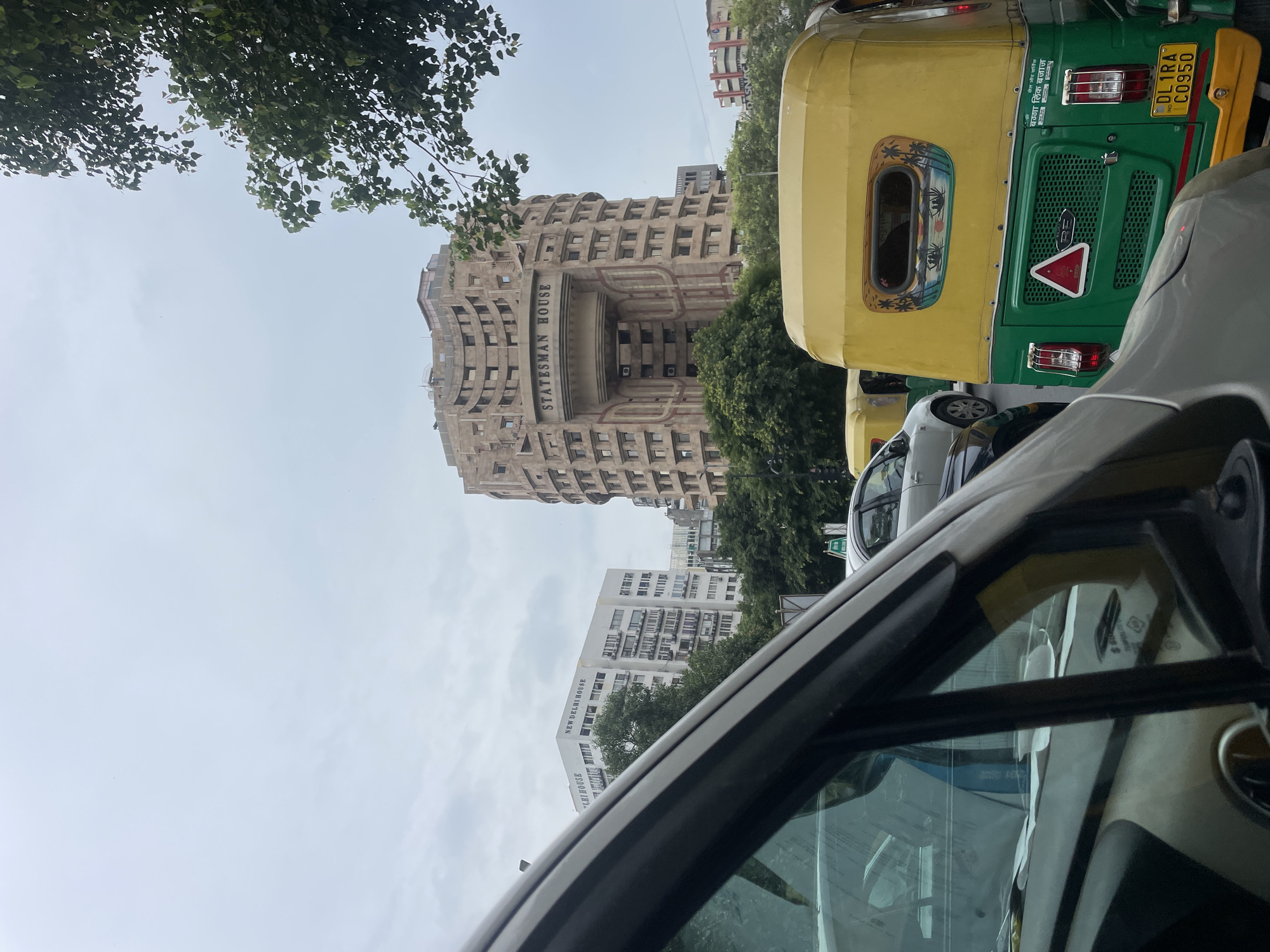 The image depicts a street scene in New Delhi, India, with a focus on the distinctive architecture of Statesman House, a well-known Brutalist building. In the foreground, the side and windshield of a dark-colored vehicle are visible, partially obscuring the view. To the right, a yellow and green auto-rickshaw is prominent, with its license plate reading "DL 1RA C0950". Behind the auto-rickshaw, other vehicles, including a white car, are in traffic. The middle ground features the curved, multi-story facade of Statesman House, characterized by its repetitive window patterns and a large, arched entranceway with the name "STATESMAN HOUSE" inscribed above it. To the left of Statesman House, a white building labeled "NEW DELHI HOUSE" is visible. Lush green trees are scattered throughout the scene, suggesting a city environment with established foliage. The sky is overcast with a pale, diffused light. The overall impression is a busy urban thoroughfare in New Delhi, with significant architectural landmarks and common modes of transportation.