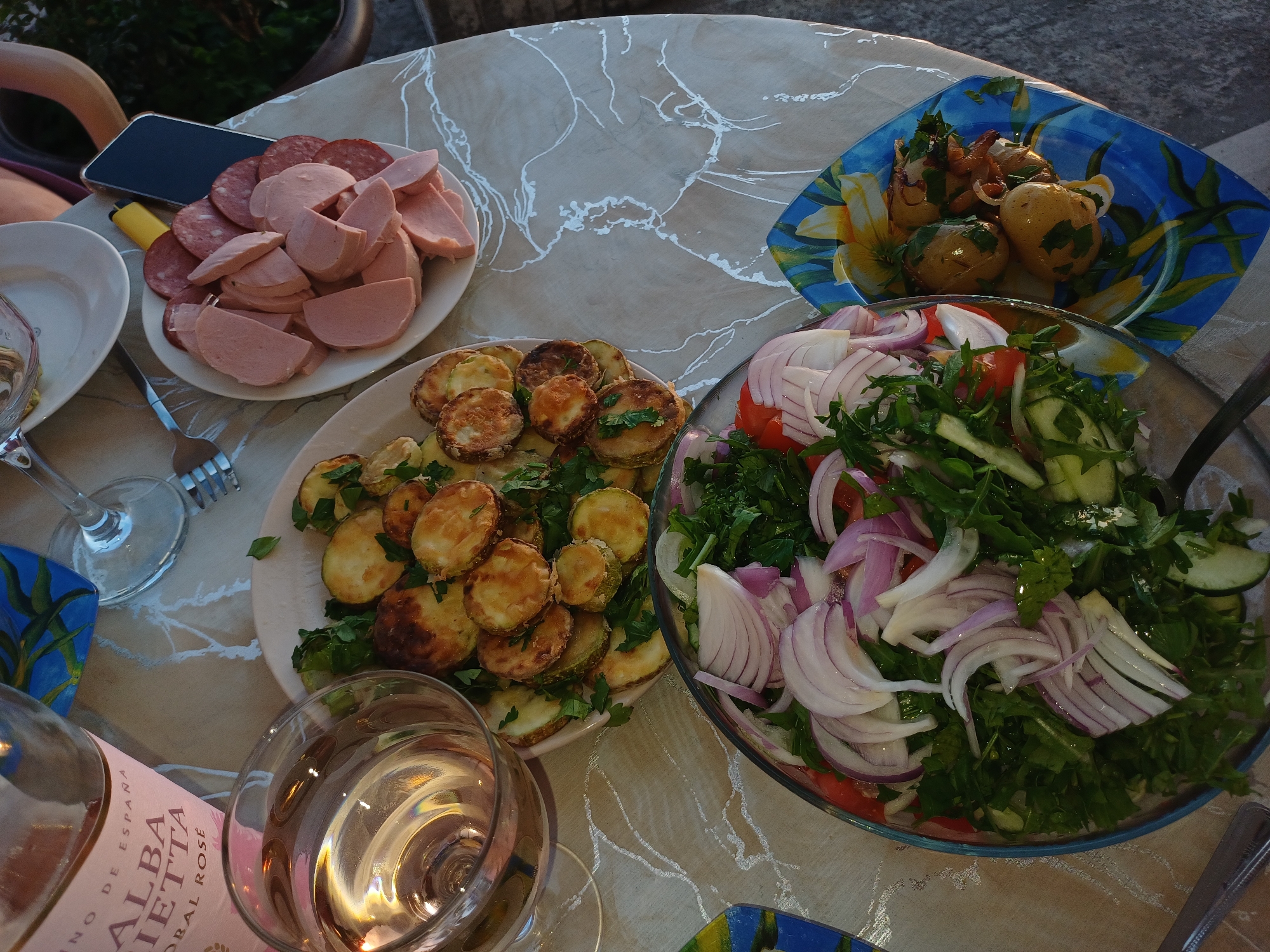 A top-down view depicts an outdoor dining table setting in Sochi, Russia, during what appears to be late afternoon or early evening. The table is covered with a light-colored surface featuring a subtle, abstract white pattern.
In the upper left quadrant, a bottle of "ALBA RIETTA CORAL ROSÉ" wine, labeled "VINO DE ESPAÑA," stands next to a clear stemmed wine glass filled with a pale rosé liquid. An empty stemmed wine glass and a silver-colored fork rest on the table adjacent to a small, empty white plate. A light-skinned human arm with a hand grasping a fork is partially visible in the upper right, positioned above this empty plate.
Centrally located on the table is a white plate laden with numerous golden-brown, pan-fried or roasted round slices of zucchini, generously garnished with chopped green herbs. To the right of this plate, another white, oval-shaped plate contains an arrangement of sliced deli meats: a lighter pink, thicker-cut variety, and a darker red, speckled, thinner-cut variety. A dark-colored smartphone stands upright on this plate, leaning against the meat slices, with a yellow disposable lighter positioned beside it.
In the lower left, a large, clear glass bowl holds a mixed salad. The salad's visible components include thinly sliced red onions, green leafy vegetables, chopped cucumbers, and diced tomatoes, all topped with fresh green herbs. Finally, in the lower right, a distinct rectangular blue plate adorned with yellow and green floral motifs displays several small, whole, cooked potatoes, garnished with herbs and what appears to be caramelized onions or other sautéed vegetables. The immediate background shows green foliage, and the ground in the lower right consists of concrete or asphalt.