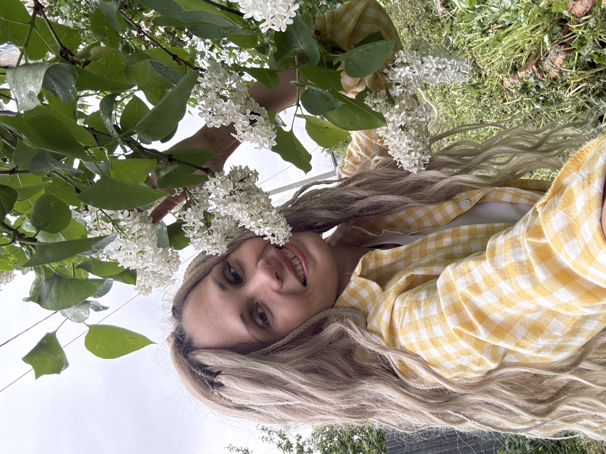 A person is standing outdoors in Rusanovo, Russia near a white lilac tree. The person has long, wavy, light-colored hair and is wearing a yellow-and-white checkered shirt. The person is smiling and holding a branch of the lilac tree. The tree has green leaves and white flowers. The ground is covered in grass and foliage. Power lines are visible in the background. The sky appears overcast.