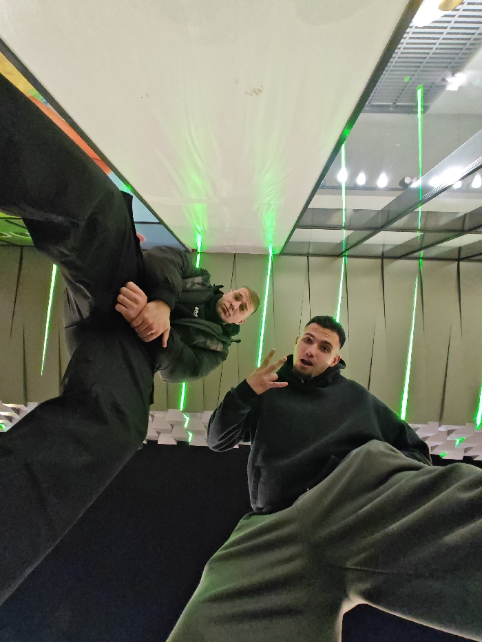 The image displays two males posing within an indoor entertainment facility identified as LaserLand Shumen, Bulgaria. The photograph is taken from a low angle, looking upwards, which distorts the perspective and emphasizes the subjects' lower bodies.
The male on the left, with short hair and a light complexion, is dressed in a dark puffer jacket over a dark top and dark pants. His legs are positioned prominently towards the camera, while his hands are clasped together at chest level. He maintains direct eye contact with the camera, exhibiting a neutral facial expression.
The male on the right, possessing short dark hair and a darker complexion, wears a dark hooded sweatshirt and grey sweatpants. His legs are also extended towards the camera. He forms a peace sign with his right hand and looks at the camera with an open-mouthed expression.
The immediate environment consists of dark walls. A section to the right features a three-dimensional pattern of white, geometric panels. The scene is illuminated by multiple vertical green laser beams, which project upwards and reflect off a large, flat, light-colored overhead surface, likely a ceiling or a large panel. Additional overhead light sources are visible, casting reflections on this surface. The ground beneath the subjects appears dark and padded.