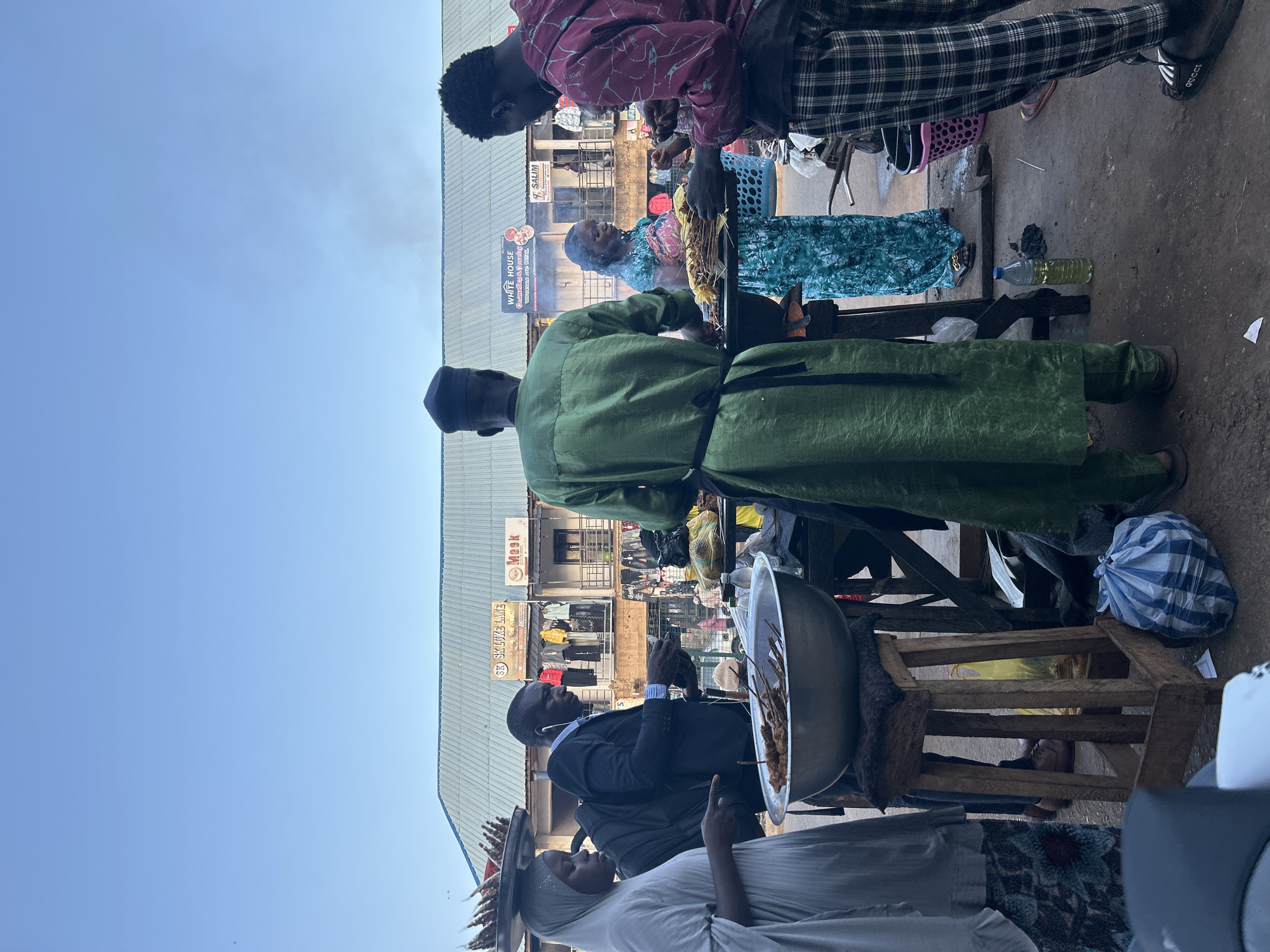 The scene captures a bustling street in Jos, Nigeria. Several individuals are engaged in various activities around what appears to be a food stall. A man wearing a green robe and a black cap is standing behind a cooking setup. To his left, another man, clad in a black suit, is interacting with a customer who is wearing a light-colored hijab, and a silver platter of cooked food. In the background, other people can be seen. The area features storefronts with signs and advertisements, and the sky is lightly overcast. The items on the cooking setup include food in baskets and a bottle of oil.
