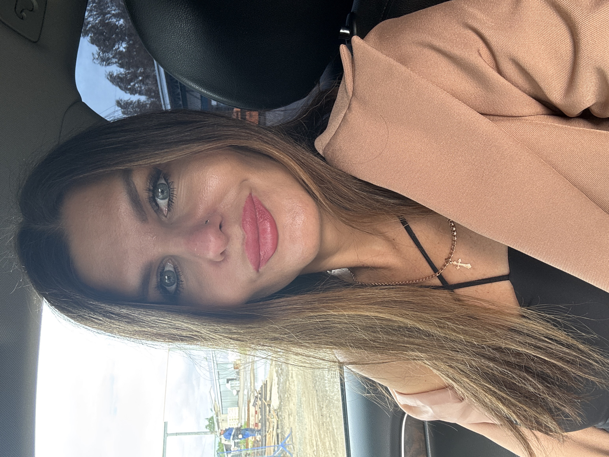 The image is a high-angle selfie of a woman, taken inside a vehicle. The woman has long, straight brown hair that flows across her shoulder and down the side of the frame. Her captivating green-blue-grey eyes look directly at the camera with a serene, slight smile. She has full pink lips and a small nose piercing. She is wearing a black top with thin straps, a gold chain necklace featuring a small cross pendant, and a light brown or tan blazer/jacket.
The setting appears to be the interior of a car, indicated by visible headrests and upholstery. Through the window on the left side of the image, an outdoor scene unfolds, revealing what looks like an industrial or construction area with buildings, utility poles, and possibly machinery, all under a bright, partly cloudy daytime sky. On the opposite side, hints of trees against a blue sky can be seen.
The woman is posing for the photograph, likely a selfie, while seated in the car. The lighting suggests it is daytime. Her expression is calm and composed. No visible text is present in the image. The location, as per context, is Chaikovsky, Russia.