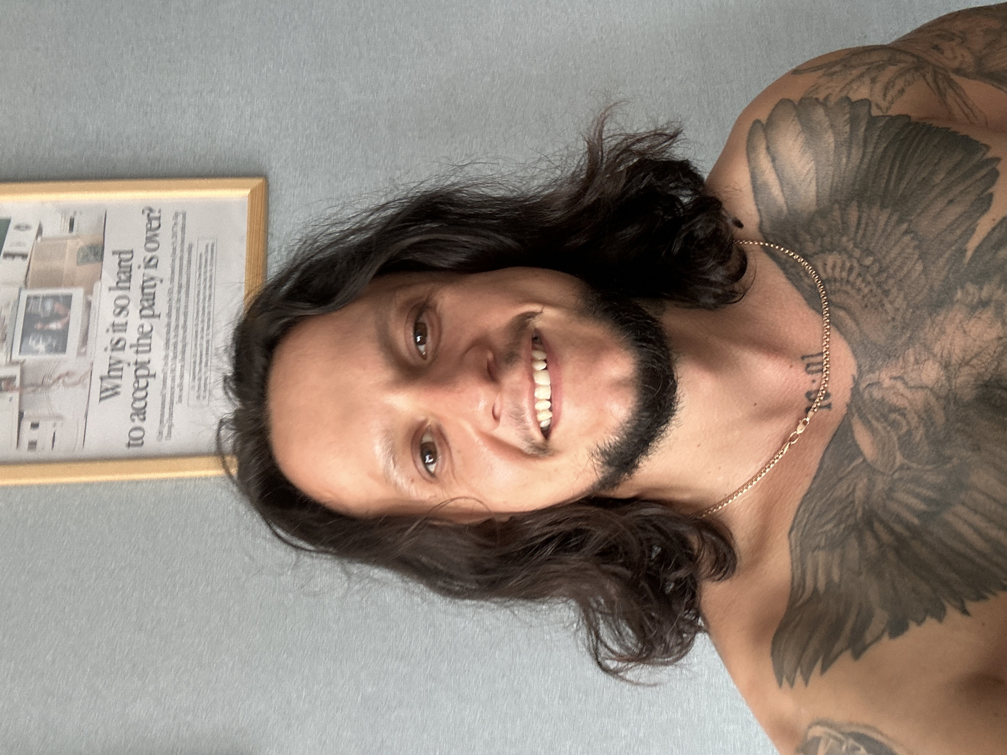 A man with long, dark, wavy hair and a goatee smiles at the camera, revealing a gold chain around his neck and a large, dark tattoo of wings covering his chest and shoulders. He is shirtless. Behind him, a framed newspaper clipping hangs on a textured, light blue wall. The newspaper headline reads "Why is it so hard to accept the party is over?". The overall environment appears to be indoors.