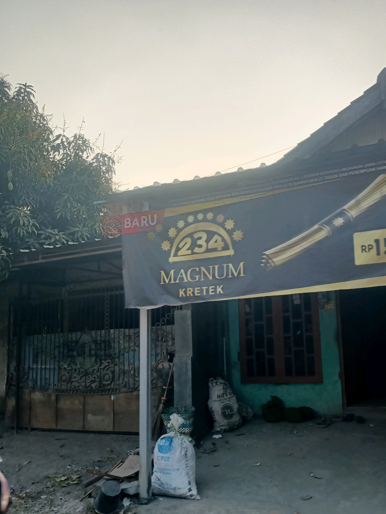 The image depicts a street scene in Pecabean, Indonesia. In the foreground, there is a paved area with scattered leaves and debris. A black bucket is visible to the left. To the right, a white sack with blue lettering and a broom leaning against it are present. Beyond this, there is a grey metal gate with ornate detailing.
The main subject of the image is a large banner hanging on the front of a building. The banner is predominantly black and gold, featuring text that appears to advertise "234 Magnum Kretek" and includes the word "BARU" (new). A portion of a golden cigar or cigarette is also depicted on the banner, along with the price "RP 15".
The building itself has a light-colored roof and walls that appear to be painted green. A wooden-framed window with multiple panes is visible on the right side of the building. To the left of the building, a tree with green leaves and what appear to be fruits is present. The sky is overcast and bright, suggesting it may be daytime. The overall impression is of a commercial establishment in a residential area.