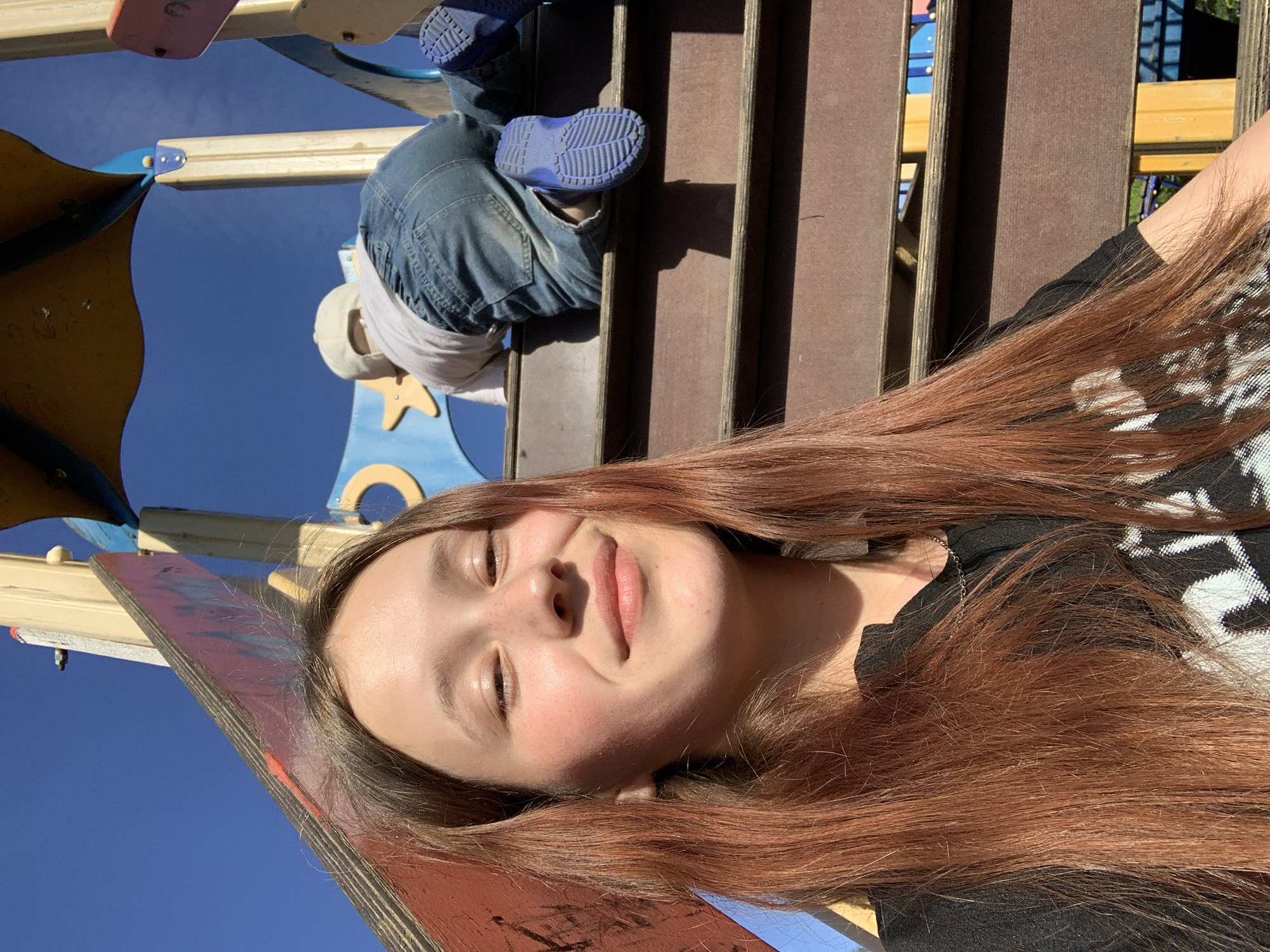 A girl is lying on a red and blue plank, possibly part of playground equipment, with her head tilted towards the camera. She has long, reddish-brown hair and is wearing a black patterned shirt. In the background, there is a child on a wooden structure with blue shoes and jeans. The overall setting appears to be a playground under a bright blue sky. The location is Фоки, Russia.