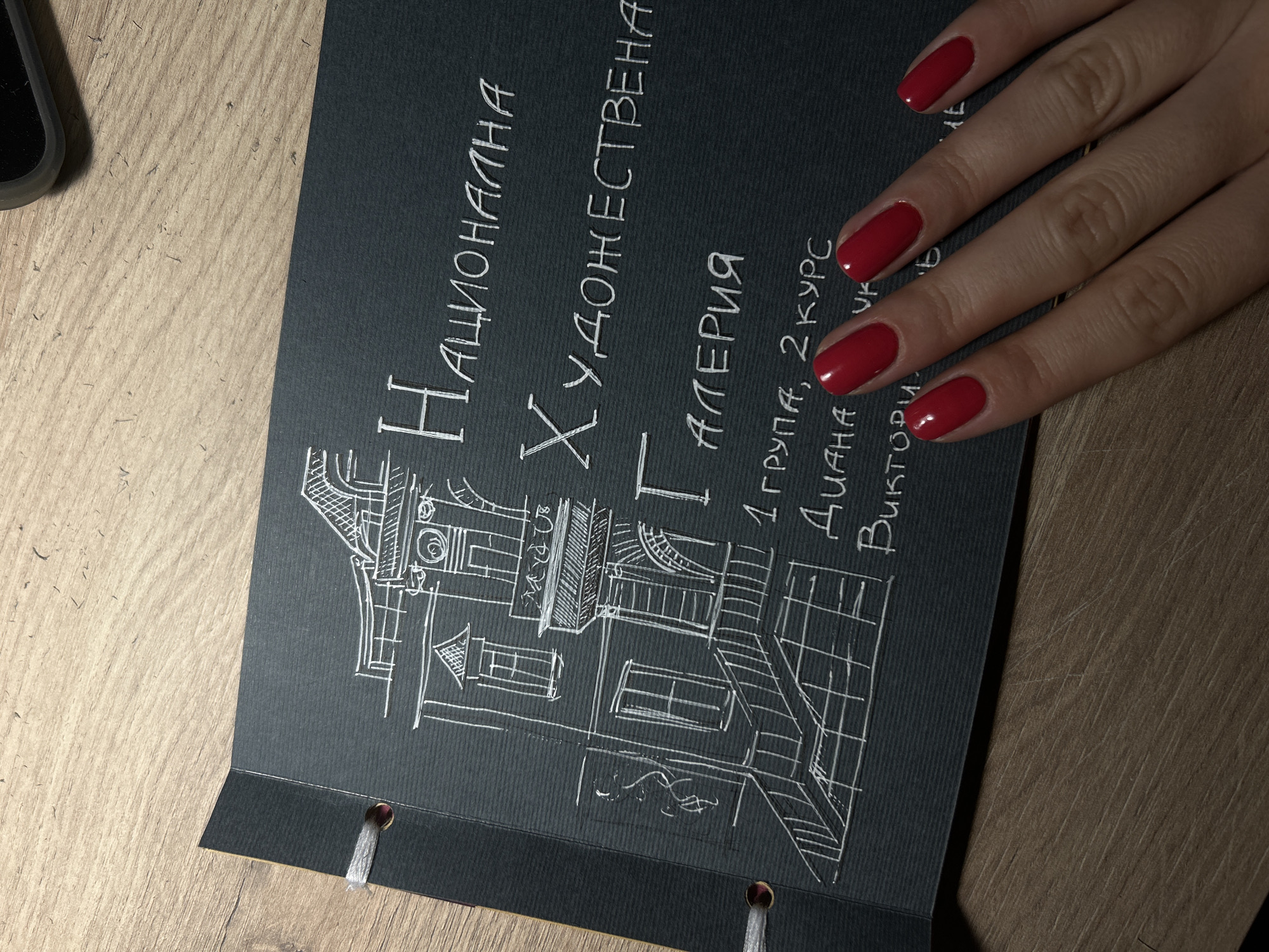 The image displays a close-up view of a dark-colored page, possibly from a scrapbook or portfolio, laid on a wooden surface. White ink outlines a drawing of a building facade, featuring windows, a balcony, and architectural details. Text in the same white ink is visible on the page. The text, written in Cyrillic script, appears to include "НАЦИОНАЛНА ХУДОЖЕСТВЕНА ГАЛЕРИЯ", which translates to "NATIONAL ART GALLERY". Below this title, further text is partially visible, reading "1 ГРУПА, 2 КУРС" (1st Group, 2nd Course) and names including "ДИАНА" and "ВИКТОРИЯ". A person's hand with bright red fingernails is positioned over the lower right portion of the page, obscuring some of the text. The background is a close-up of a wooden table with visible grain. The scene is lit from above, creating subtle shadows.