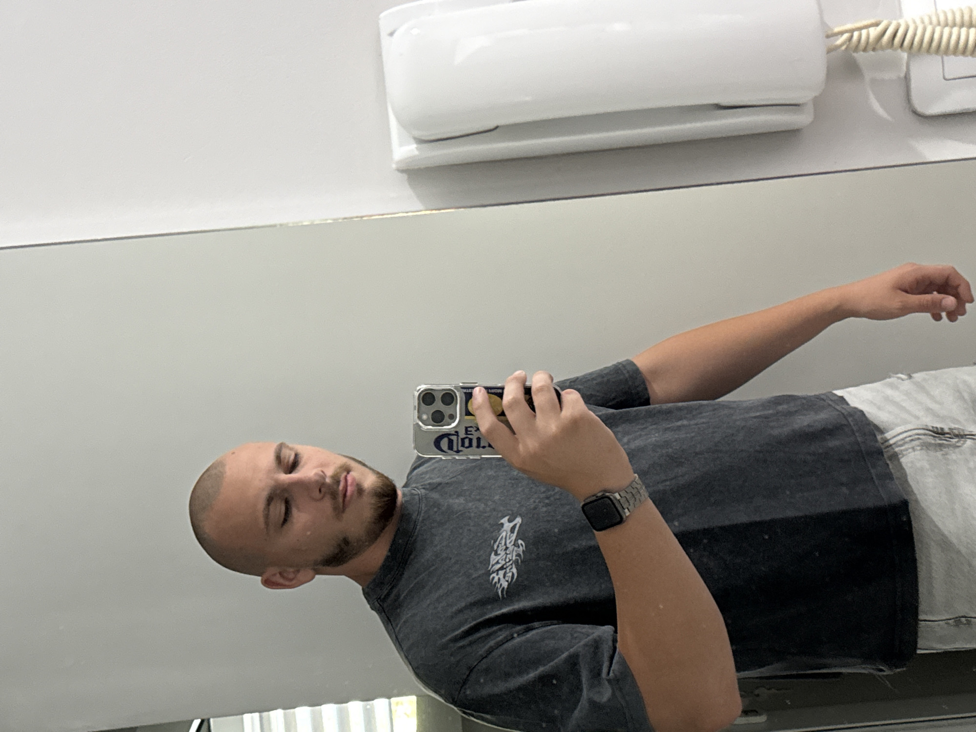 A young man with a shaved head and beard is taking a mirror selfie in what appears to be a hotel bathroom. He is wearing a dark grey t-shirt and light grey shorts. He is holding a smartphone in his right hand, which has a case with a Corona beer advertisement on it. On his left wrist, he is wearing a smartwatch. To the right of the man, a white wall-mounted intercom system is visible. The background is a plain white wall and a large mirror reflecting the man and the intercom. There is no indication of the city of Stara Zagora, Bulgaria, or any other specific location.