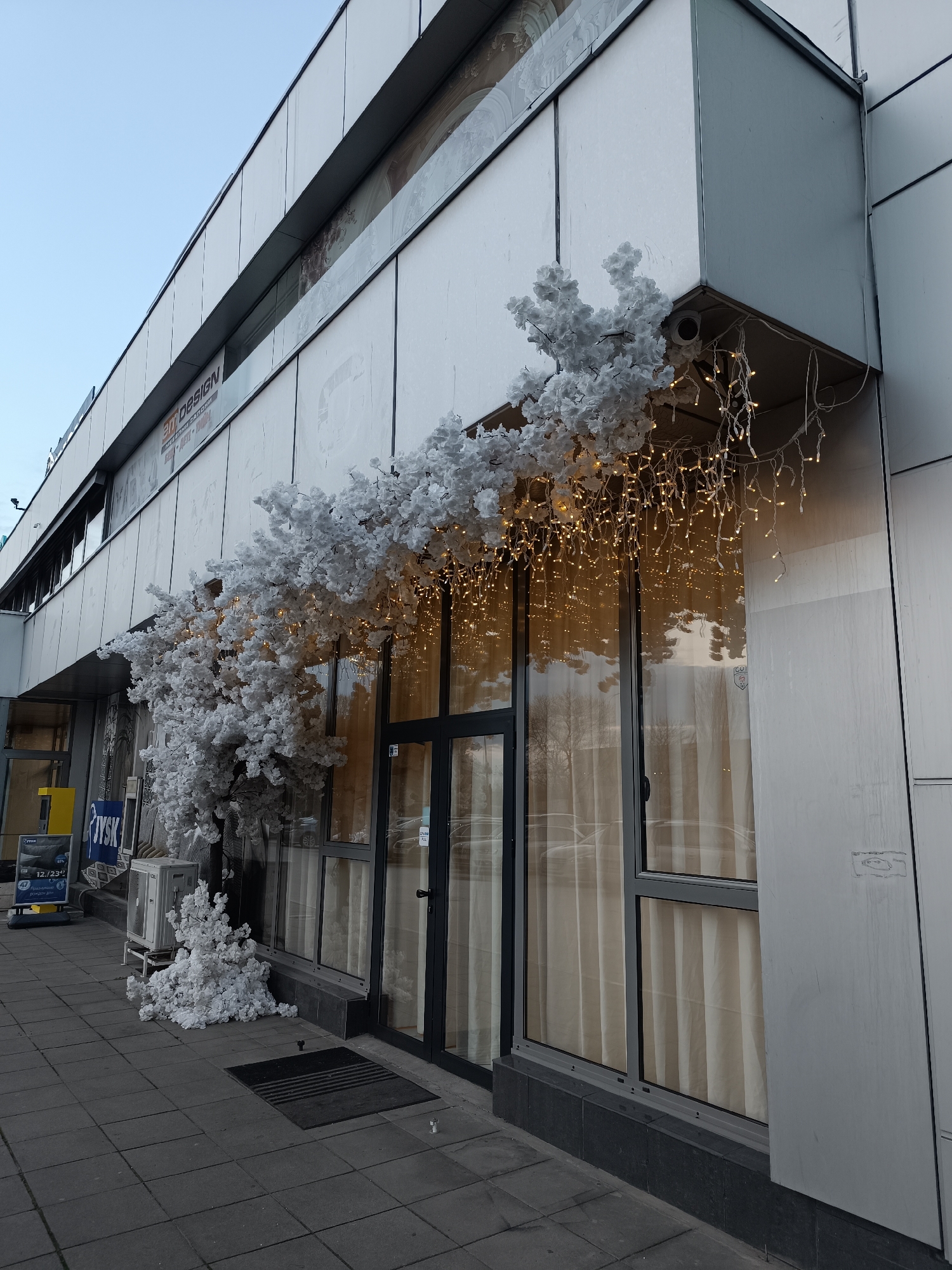 The image shows the exterior of a commercial building in Pleven, Bulgaria. The building has a modern facade with gray paneling, large glass windows, and a storefront entrance. The entrance is decorated with white artificial floral arrangements, resembling snow-covered branches, and string lights, giving a festive appearance. A "JYSK" store sign is visible to the left of the entrance. The sidewalk in front of the building is paved with gray tiles. A ventilation unit is placed near the building’s foundation. The sky is clear and light blue.