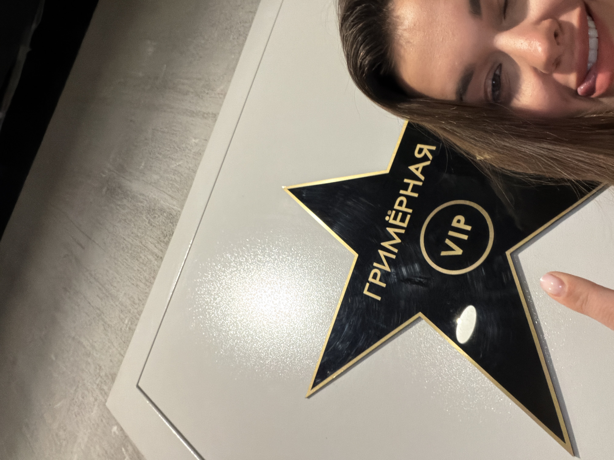 A medium shot shows a young woman winking and sticking out her tongue, positioned in the right foreground. Her right eye is closed, and her tongue is visible between her lips. She has brown hair. In the mid-ground, a black star with a gold border is affixed to a light-colored door or wall. The star has gold lettering that reads "ГРИМЁРНАЯ VIP". The background consists of a textured, grey surface, likely a wall or floor, and a sliver of a darker surface above. The overall scene appears to be indoors, possibly in a backstage or dressing room area, given the signage on the star. No specific location in Moscow, Russia, can be identified from the image.