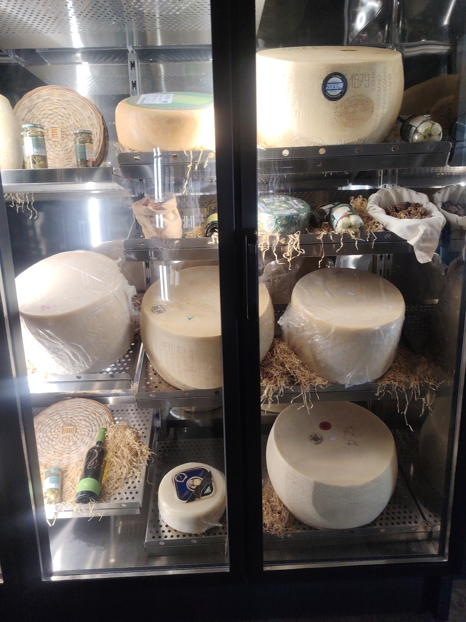 The image displays a refrigerated display case filled with various types of cheese and other food items, likely in a retail setting. The display case is well-lit, highlighting the products.
On the top shelf, a large, round wheel of cheese with a green label sits on a perforated metal rack. To its left, there's a woven basket containing two glass jars with green contents, possibly olives or pickles. Another large, pale cheese wheel is visible to the far left on the same shelf.
The middle shelf features three large, round cheese wheels. The one on the far left is covered in plastic wrap. The middle cheese wheel is also partially wrapped, and the one on the right is largely unwrapped and displays text around its circumference. In front of these cheeses, to the right, are two small cloth bags filled with walnuts and some other items.
The bottom shelf contains a woven mat on the left, next to a green bottle and a small jar. To the right, a smaller, round cheese wheel wrapped in plastic is presented on a perforated metal rack, surrounded by shredded straw-like material.
A large, oval-shaped cheese wheel, also on a perforated rack, is visible on the right side of the bottom shelf, surrounded by similar straw-like material.
The overall environment suggests a store specializing in cheeses and gourmet food items. There are no people visible in the image. The location is not Konigil, Uzbekistan, as no identifying markers or contextual clues suggest this locale.