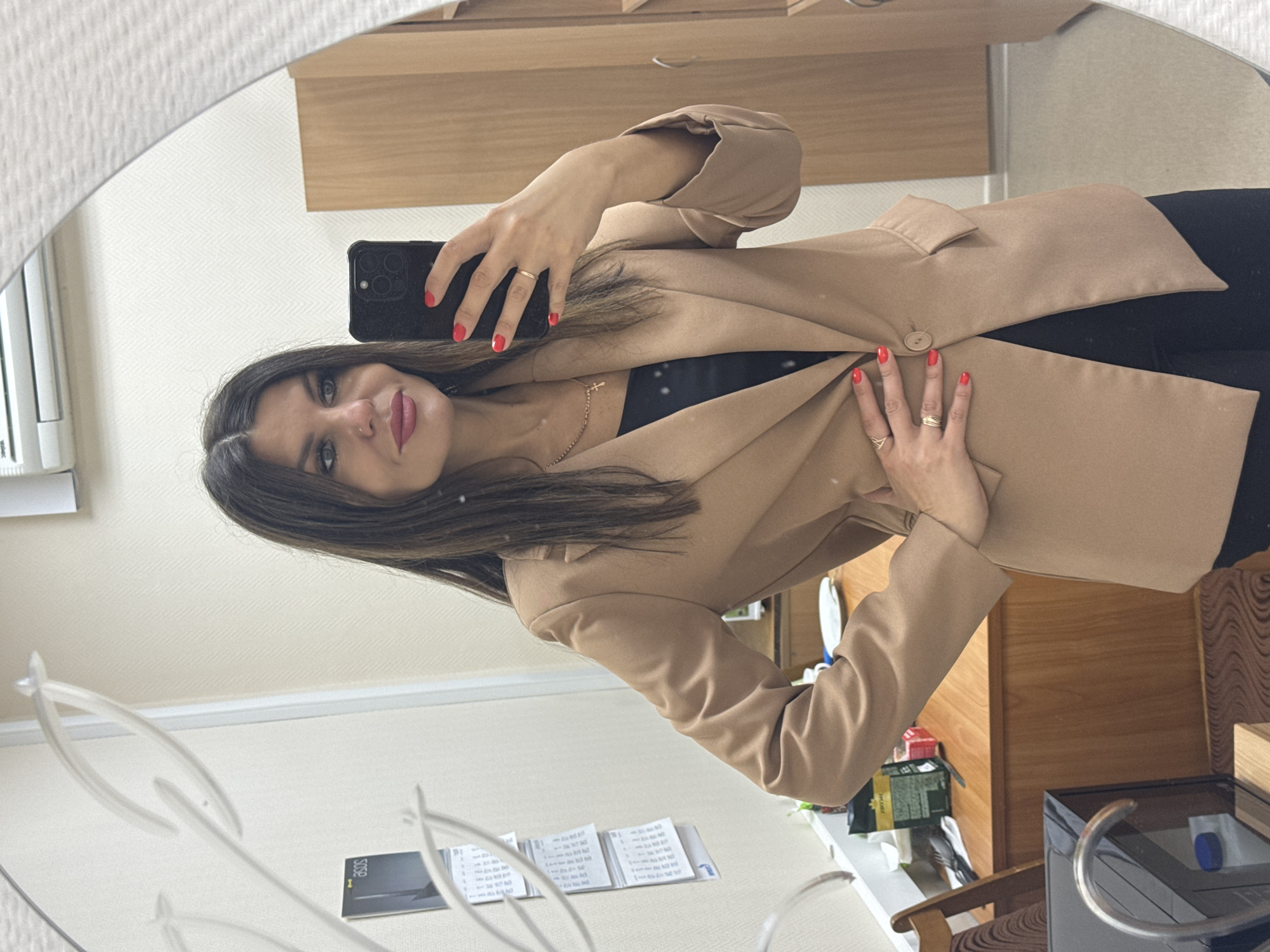 A young woman is taking a selfie in a mirror. She has dark hair and is wearing a tan blazer over a black top. She is holding a black smartphone in her left hand and has red nail polish. The mirror shows reflections of a room, including a cabinet, a desk with papers, and other miscellaneous items. The location is likely an indoor space in Chaikovsky, Russia, based on the presence of items or elements common to that region.