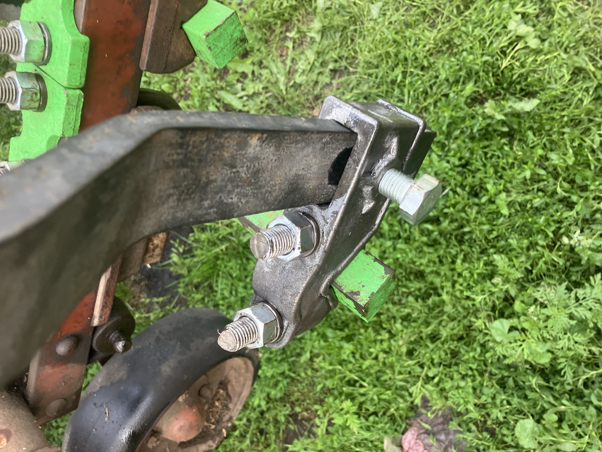 In Vovkivka, Ukraine, a close-up, low-angle shot captures the underside of agricultural equipment resting on a patch of green grass. The focal point is a heavy metal attachment with two bolts and nuts securing it. One bolt is partially obscured by the attachment, while the other is fully visible. A green, rectangular block protrudes from the bottom of the metal attachment. To the left, a black tire, likely from a wheelbarrow or small tractor, is visible against a red and orange metal frame. The background consists of a dense growth of green grass.