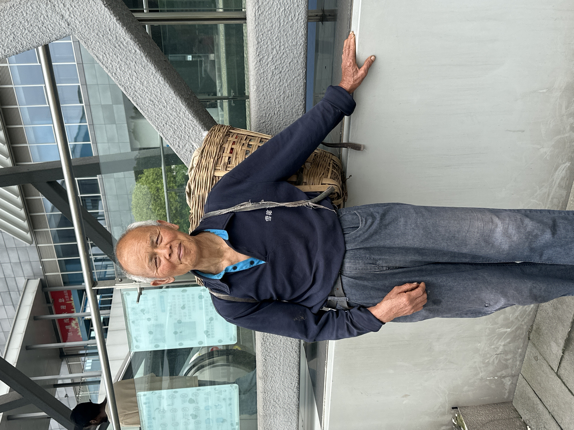 An elderly man with wrinkled skin and thin white hair is standing on a concrete surface, likely a sidewalk, with a woven basket strapped to his back. He is wearing a dark blue collared shirt with a lighter blue trim and button closure, and grey-blue denim pants. His right arm is extended and his hand is pressed against a grey wall, while his left hand is gesturing slightly near his waist. The background features a modern building with glass windows and steel supports, partially visible due to the tilted perspective. Red Chinese lettering is visible on a sign inside the building. The location is Jiangjin, China.
