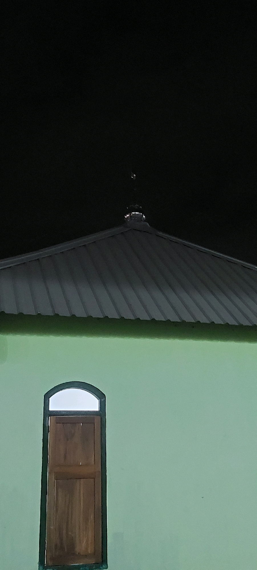This is a vertical, eye-level, medium shot of a building at night. The top of the image shows a dark sky with a peaked, dark gray metal roof that has horizontal corrugated lines. On the apex of the roof, there is a small, reflective object with what appears to be a finial. Below the roof is a band of bright green, and beneath that is a light green wall. Centered on the wall is a tall, arched window with a dark brown wooden frame and shutter. The shutter is divided into two panels by a horizontal line. The top section of the arched window is clear, showing a bright white interior. The image is dark and grainy due to the low light conditions. The location is Unknown, Unknown.