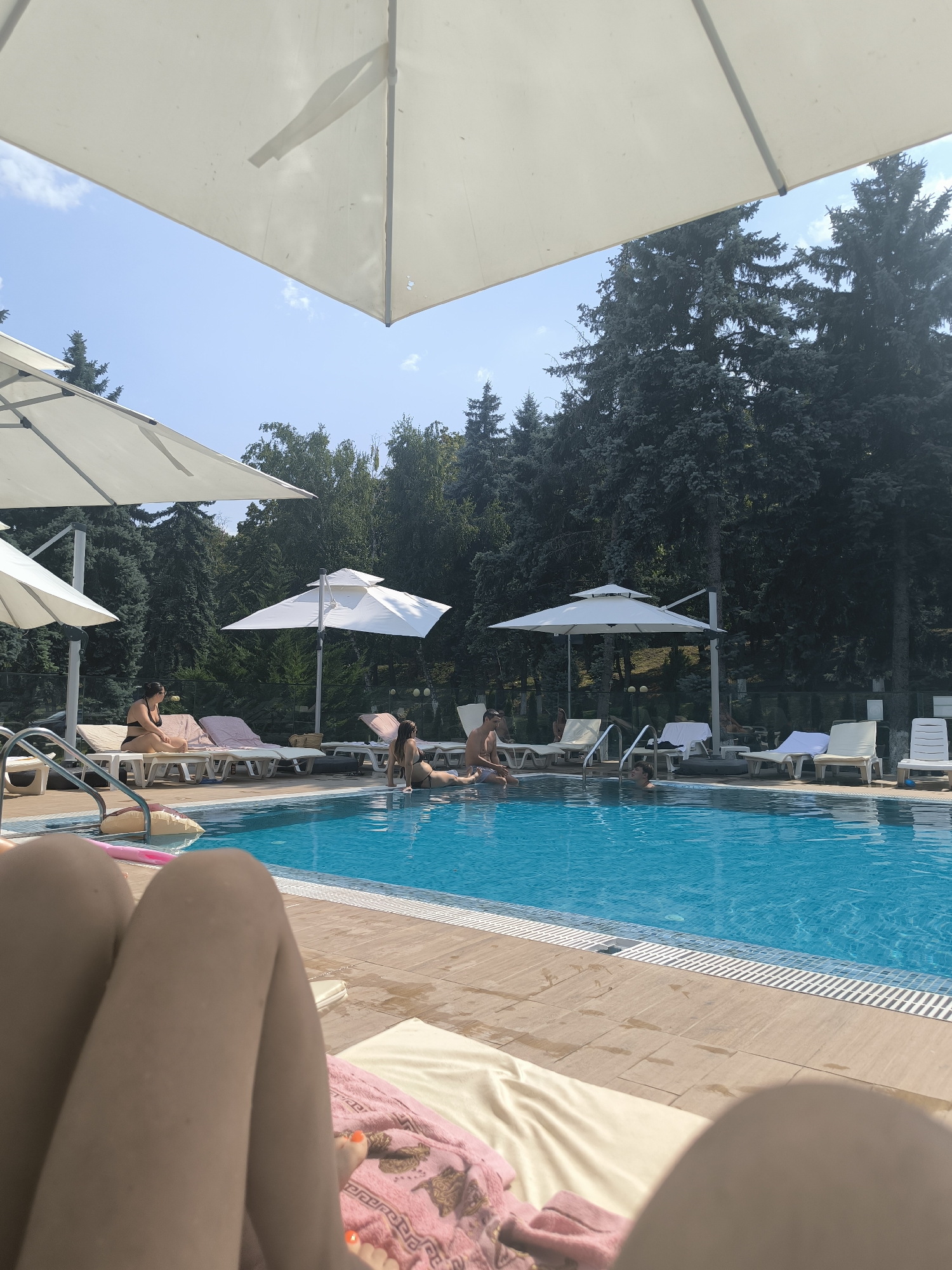 In Ustia, Moldova, a clear blue swimming pool is observed with several individuals present. The pool area is surrounded by lounge chairs and large white umbrellas providing shade. A woman in a black bikini is seated on a lounge chair on the left side of the image, facing towards the pool. Another woman, also in a bikini, is sitting at the edge of the pool, with her legs submerged in the water. Further into the pool, a man is partially visible, with his upper body out of the water. The background features a dense line of evergreen trees, suggesting a natural or park-like setting. The foreground of the image is occupied by the legs and lower torso of a person lying on a lounge chair, with an orange-painted toenail visible. The overall scene depicts a leisurely day at an outdoor swimming pool.