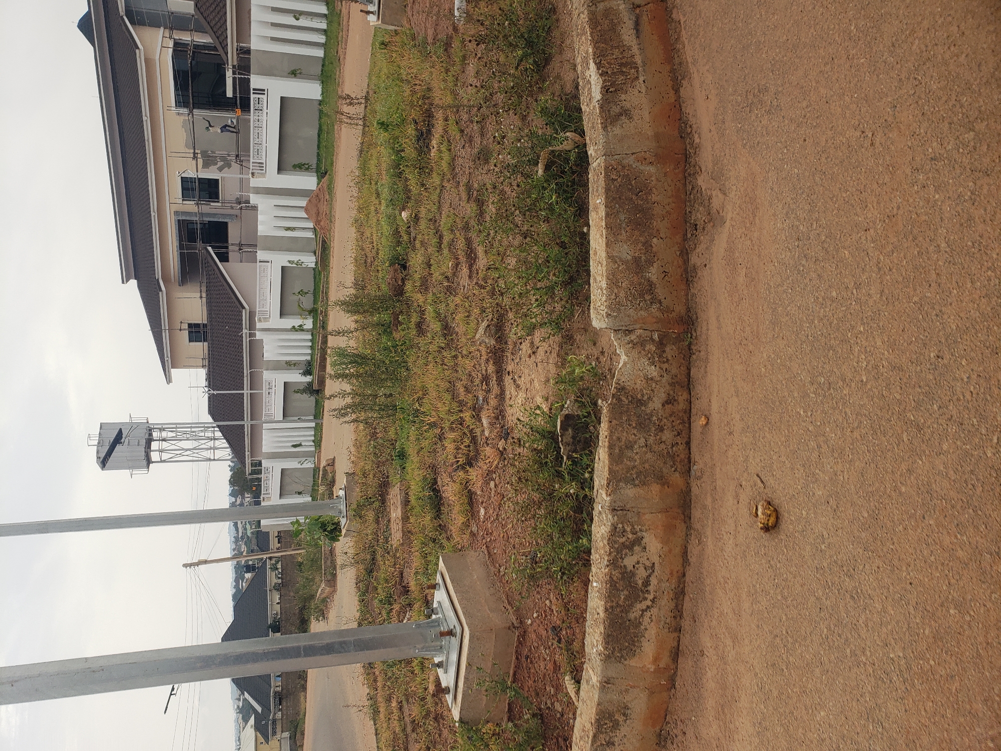 A paved road curves around a circular island of dry, sparse vegetation. Two metal poles, likely streetlights or part of an overhead electrical system, rise from concrete bases on the island. The island is bordered by a concrete curb. In the background, a large, modern house under construction is visible, with scaffolding around a section of its exterior. A water tower with a metal support structure stands behind other residential buildings further in the distance. The sky is overcast. A small pile of yellow-brown material, possibly refuse or debris, lies on the paved road in the foreground. The environment suggests a residential development in Rayfield, Nigeria, likely undergoing new construction.