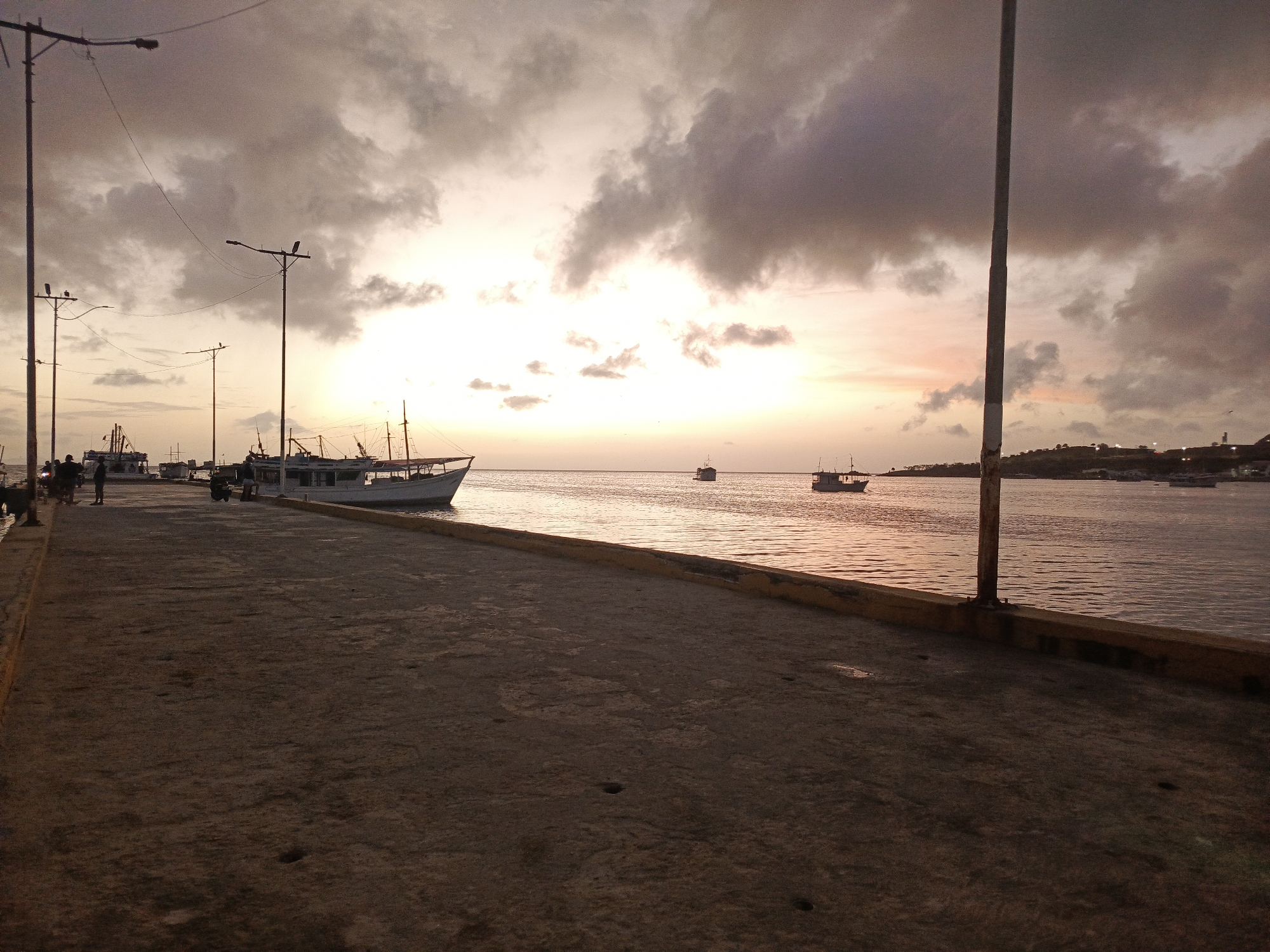 The image depicts a twilight scene at the harbor of Juan Griego, Venezuela. The sky is overcast with dramatic clouds tinged with the warm hues of sunset. Several boats, varying in size and design, are anchored or moving in the calm waters of the bay. A concrete pier or walkway occupies the foreground, with a yellow-lined edge separating it from the water.
On the pier, in the left portion of the image, are silhouetted figures of people and what appears to be a motorcycle. Streetlights on poles are visible along the pier, some with electrical wires connecting them. In the middle ground, larger boats are moored, their white hulls contrasting with the darker water. Further out in the bay, smaller boats are scattered, and in the distance, the coastline is faintly visible with structures and lights indicating a settlement. The overall atmosphere is serene, suggesting the end of the day in a coastal town.