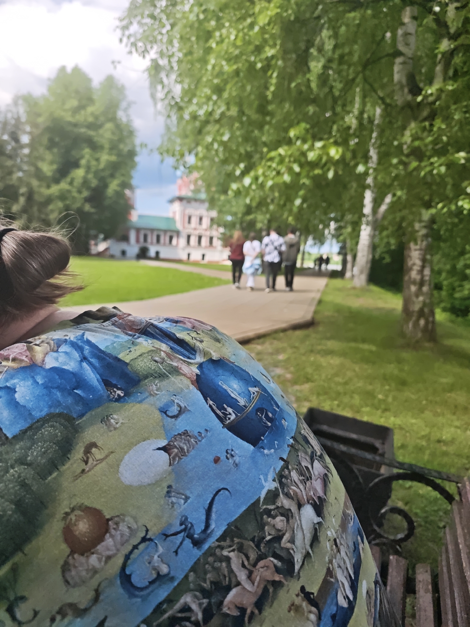 A woman with dark hair in a bun sits on a wooden bench in a park. She is wearing a shirt featuring a printed design that appears to be a collage of paintings. In the background, a group of people is walking along a paved path towards a building with red and white architectural details. Green trees and grass flank the path and the building, suggesting a park setting. The photo appears to be taken from behind the woman, focusing on her clothing and the scene beyond. The city is Selivanovo, Russia.