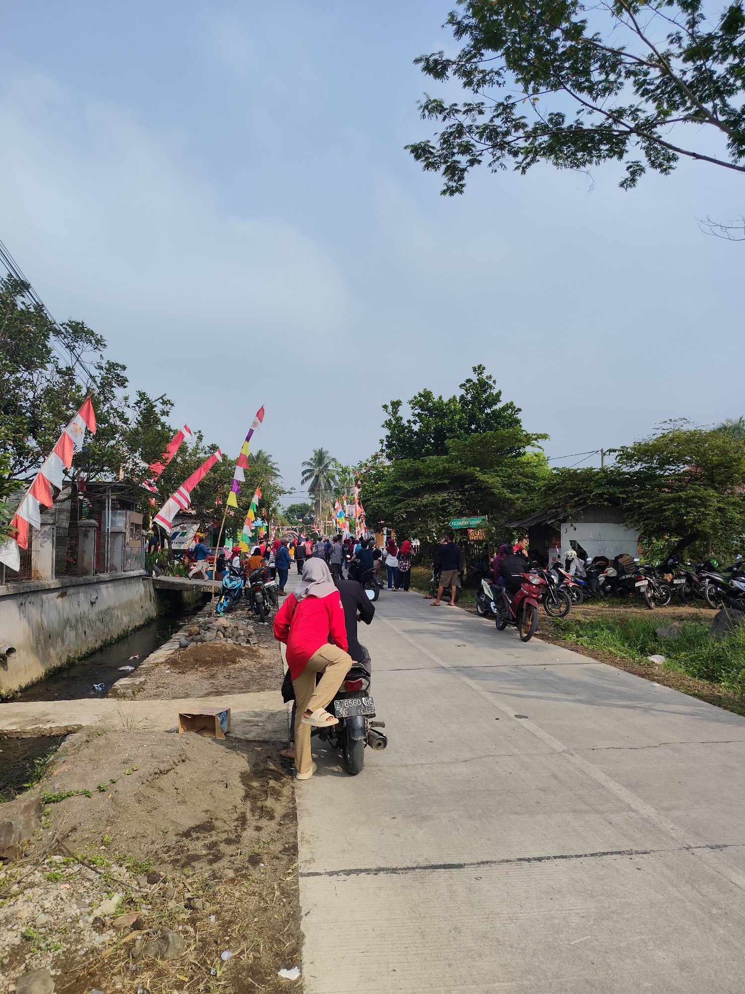 This is an outdoor, eye-level, full shot of a street scene in Sukamahi, Indonesia, likely during a celebration or event. The sky is clear with scattered clouds, and trees line the right side of the road. Many red and white Indonesian flags are strung along the left side of the street, some on poles and some attached to wires. A narrow concrete canal runs parallel to the left side of the road, with debris visible in the water.

In the foreground, a person wearing a red shirt and tan pants is seated on a black motorcycle. Their left leg is bent, and their foot rests on the pavement. Another person wearing a gray hijab is also visible behind the rider. The motorcycle's license plate reads "Z 6560 HL 06-22".

The road itself is paved with concrete and marked with white lines. A crowd of people and motorcycles are visible further down the street. To the right of the road, there are more trees and some small buildings, one of which appears to be a food stall. Several motorcycles are parked along the side of the road. The overall impression is a lively, community gathering in a semi-urban or rural Indonesian setting.
